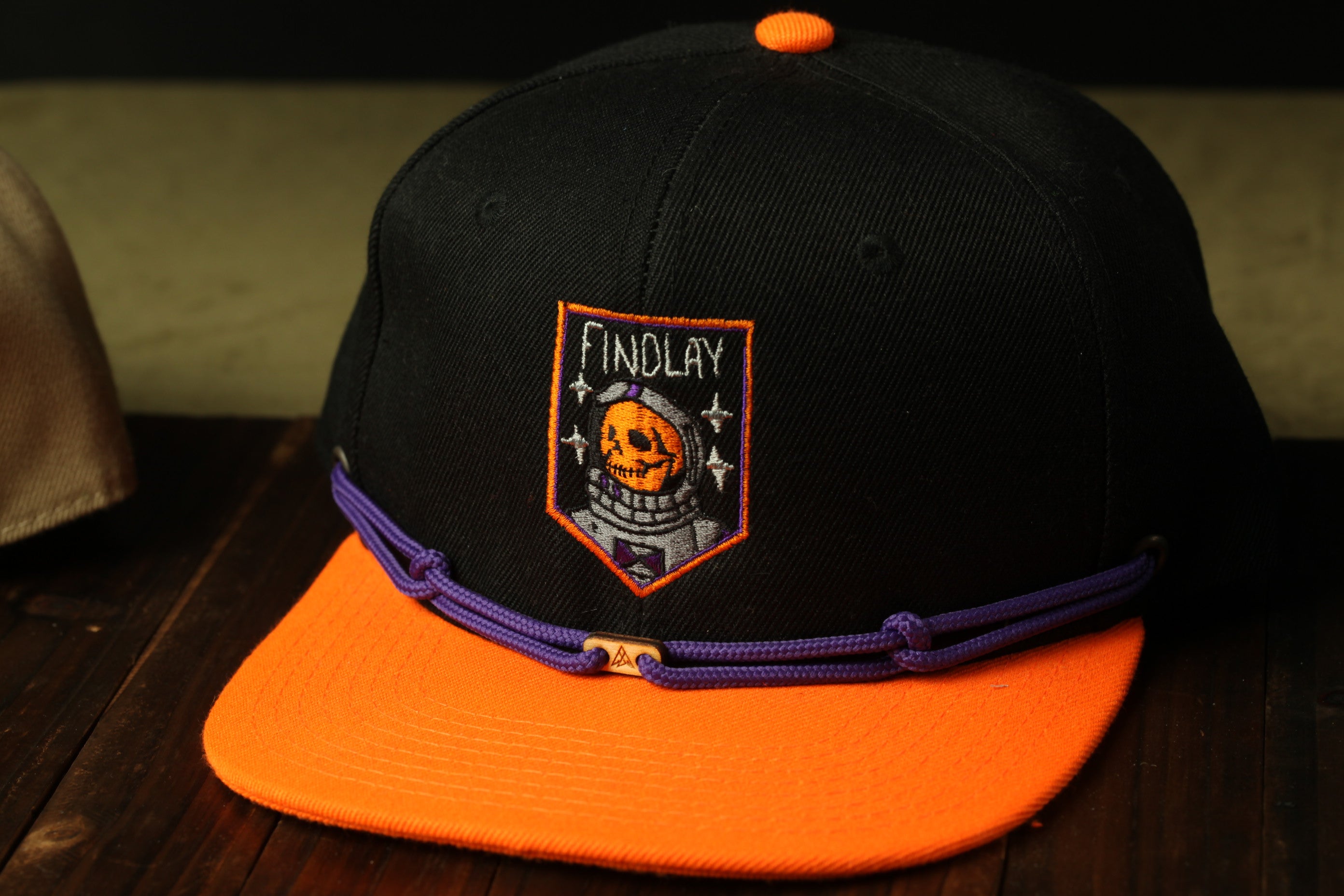 A black baseball cap with an orange brim and a purple band around the brim, featuring an embroidered patch with a skull and the word "FINDLAY" on it.