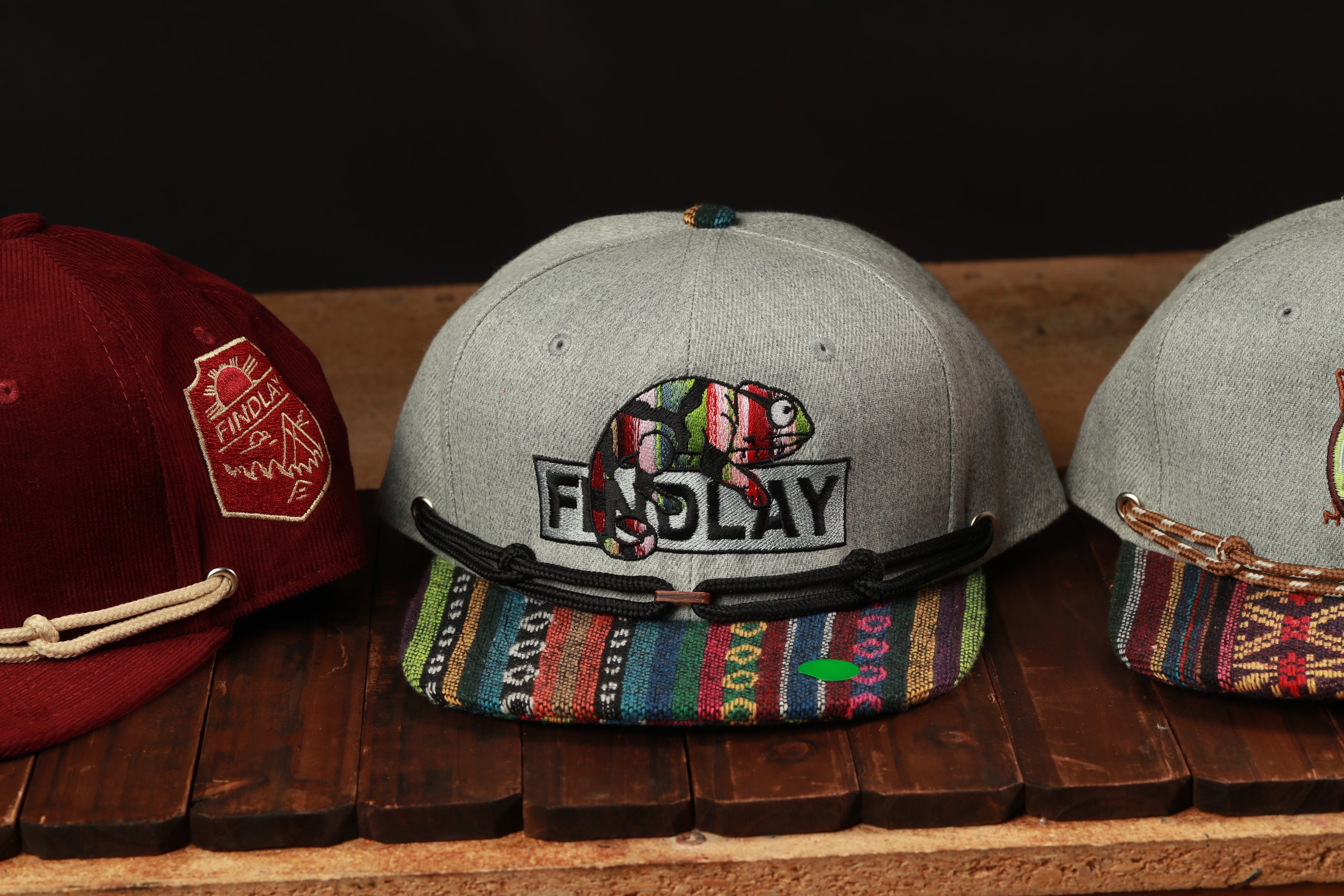 Three baseball caps are displayed on a wooden surface, with one gray cap featuring a colorful pattern and the word "FINDlay" printed on it.