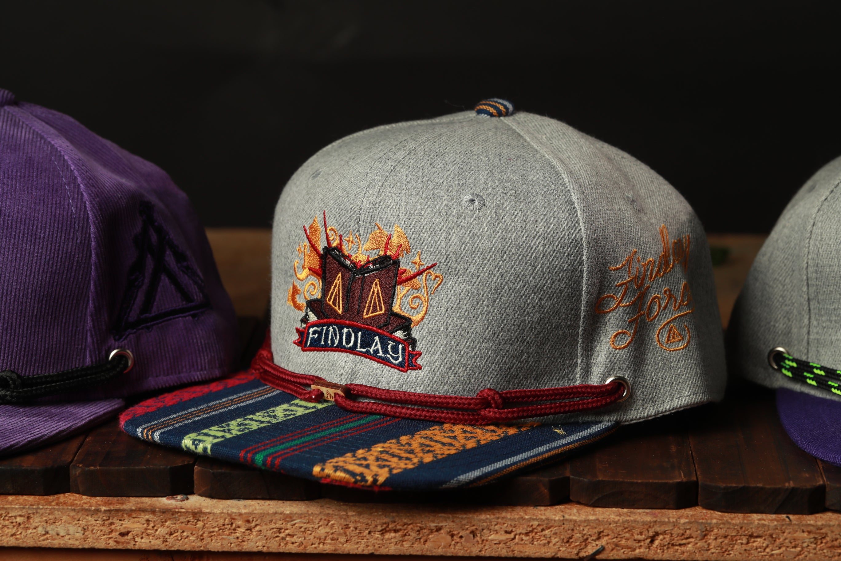A gray baseball cap with a colorful, embroidered design and the word "FINDlay" printed on it, along with a red string and a purple cap.