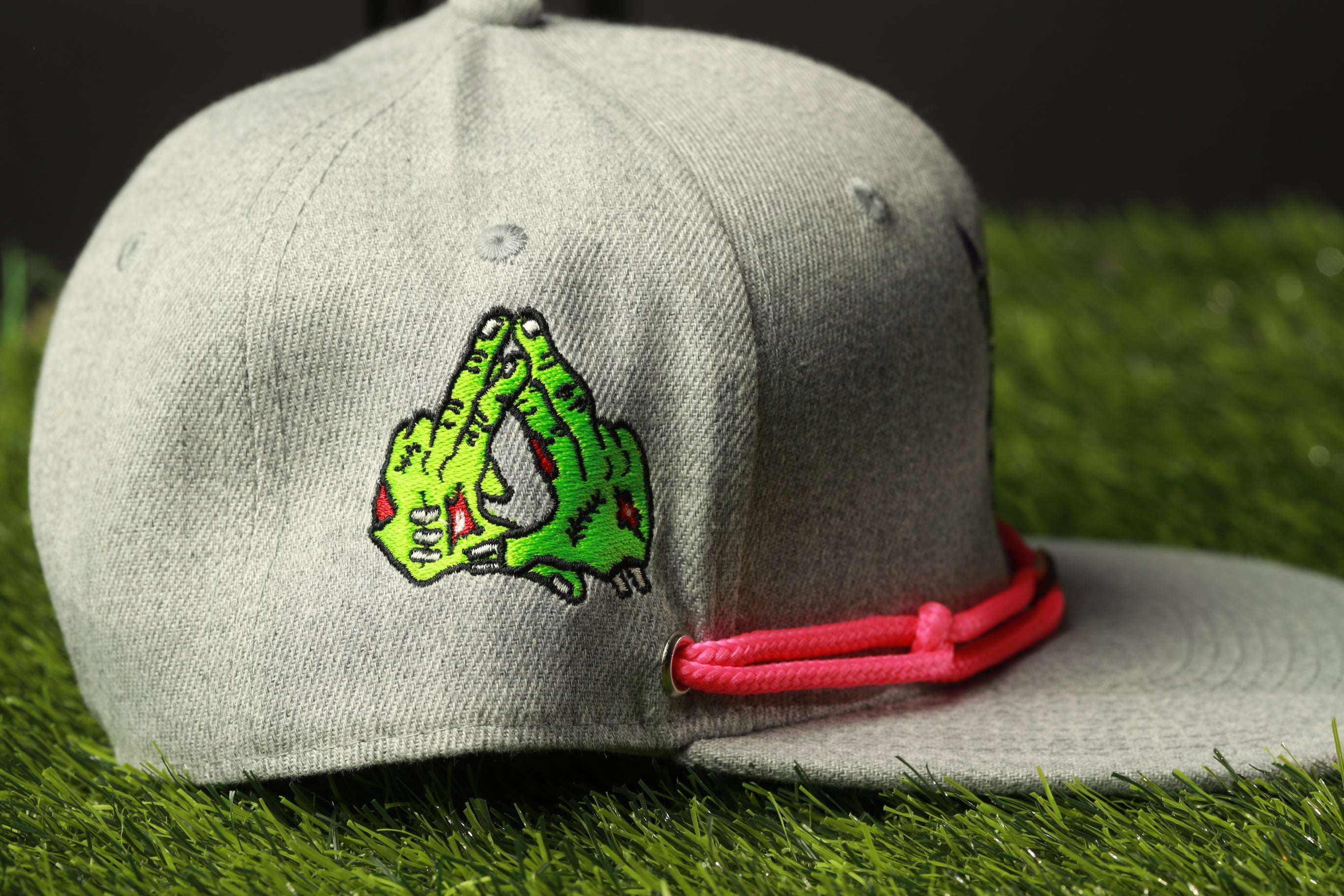 A gray baseball cap with a green and pink design on the front, placed on a grassy surface.