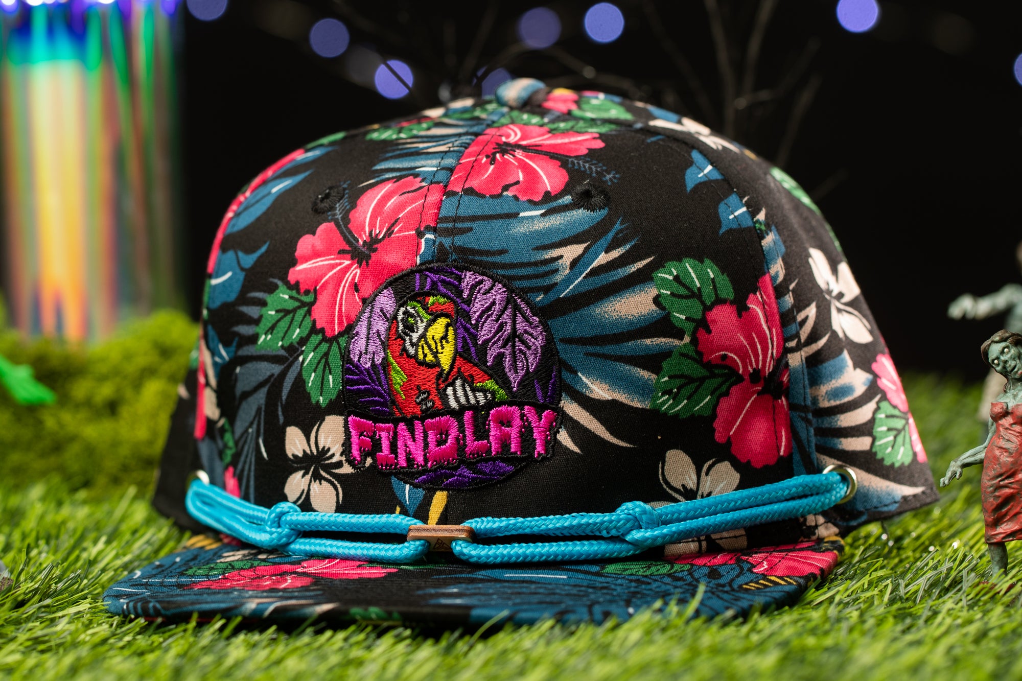 A colorful floral baseball cap with a bird embroidered on it, placed on a grassy surface with a blurred background.
