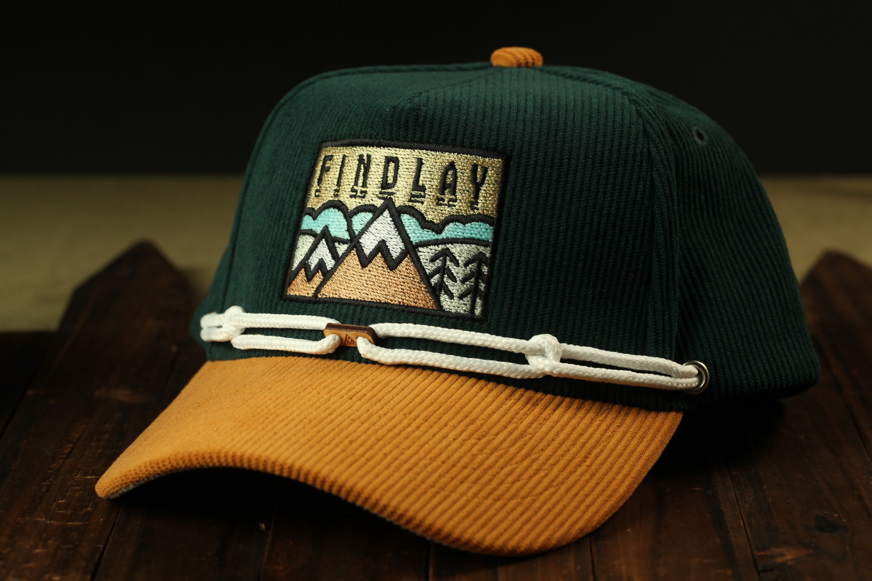 A green baseball cap with a mountain-themed patch and a white string, placed on a wooden surface.