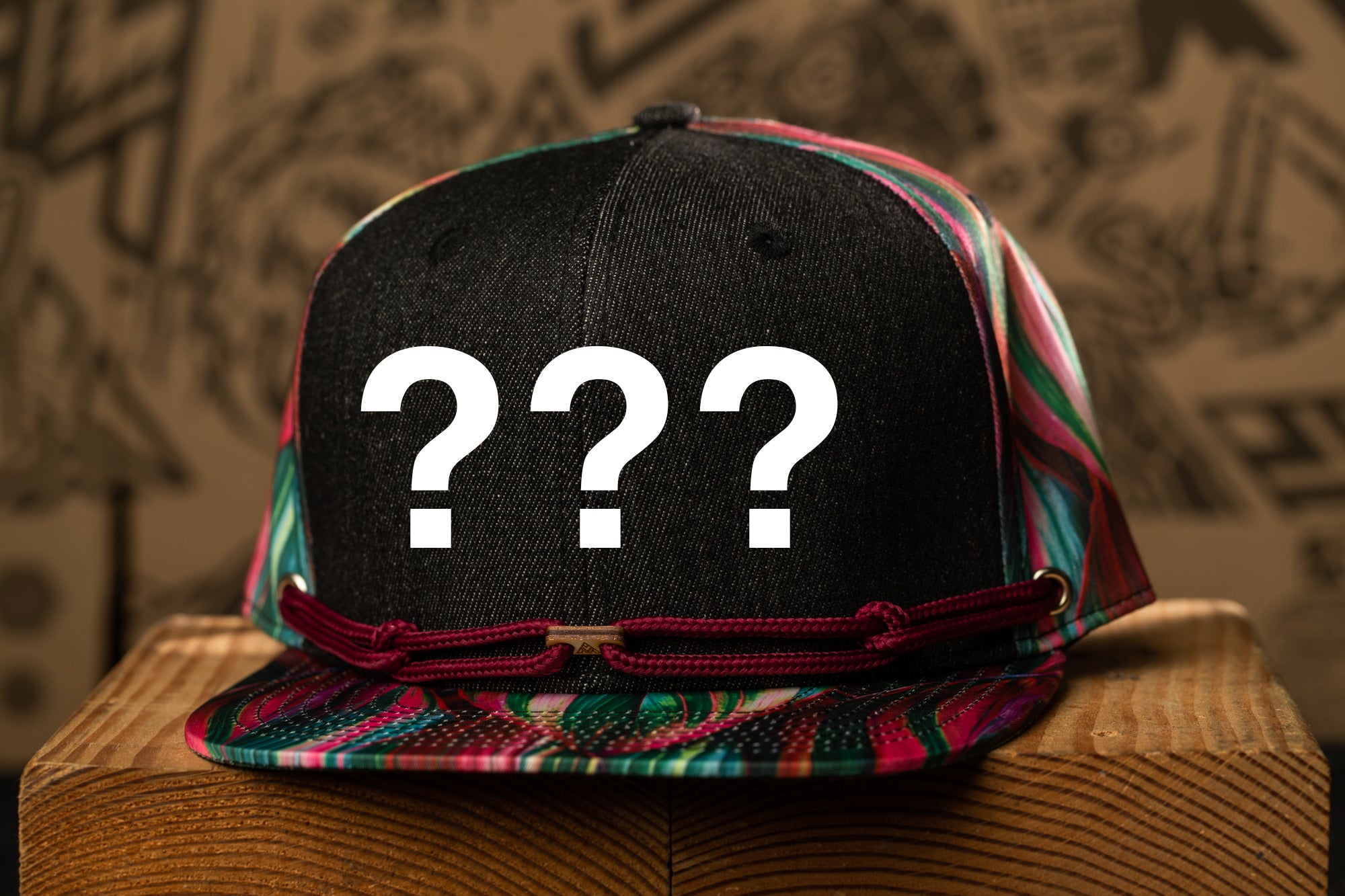 A black baseball cap with a colorful pattern and three white question marks on it, placed on a wooden surface against a background with abstract designs.