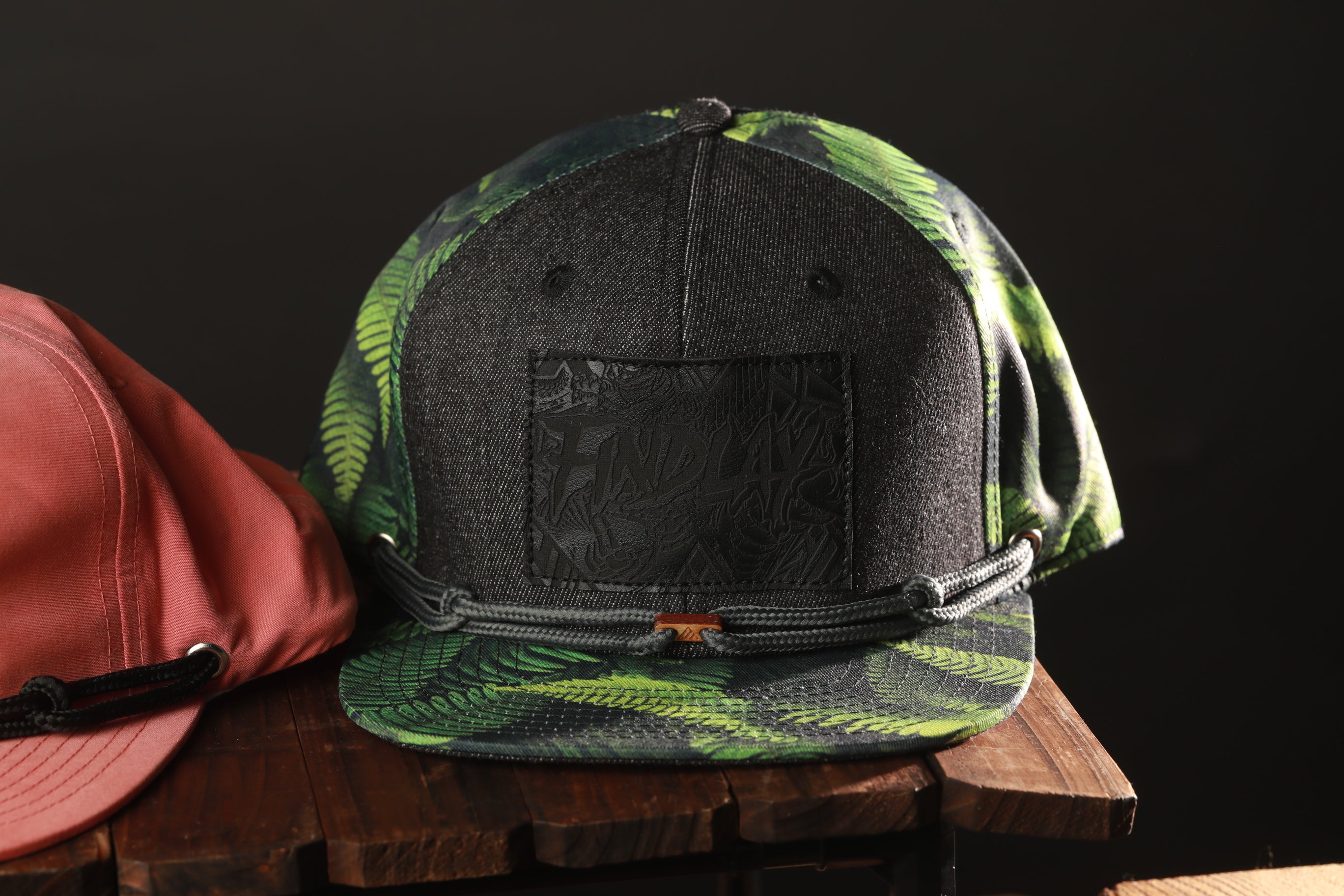 A black and green camouflage baseball cap with a black band is displayed on a wooden surface, next to a red baseball cap.