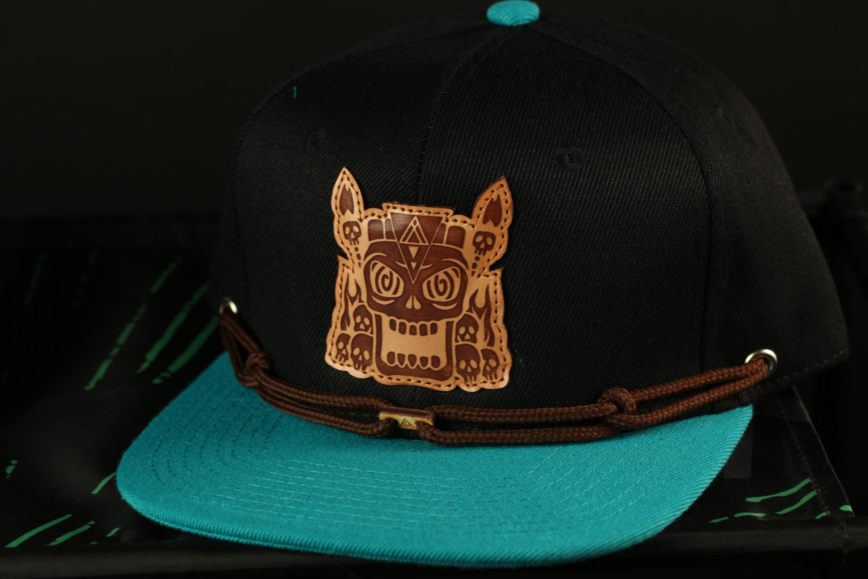 A black baseball cap with a teal brim and a brown leather-like design on the front.