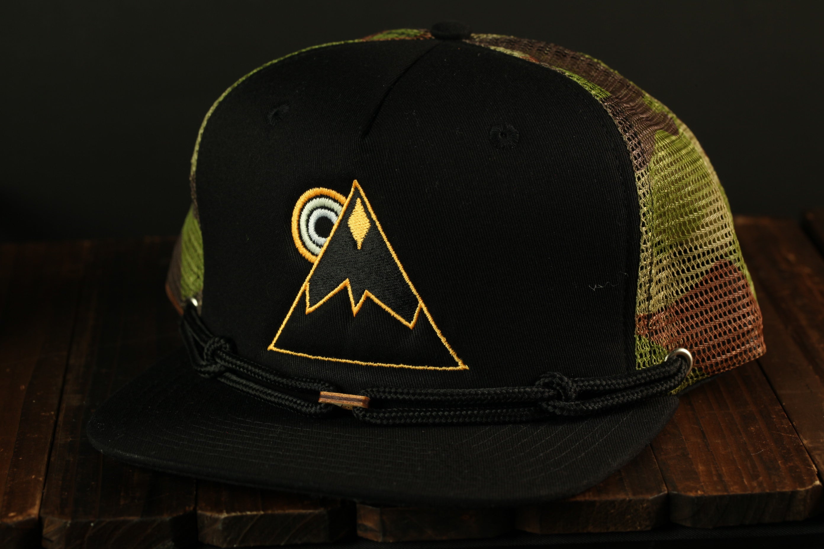 A black baseball cap with a camouflage pattern and a yellow triangle design on the front, sitting on a wooden surface.