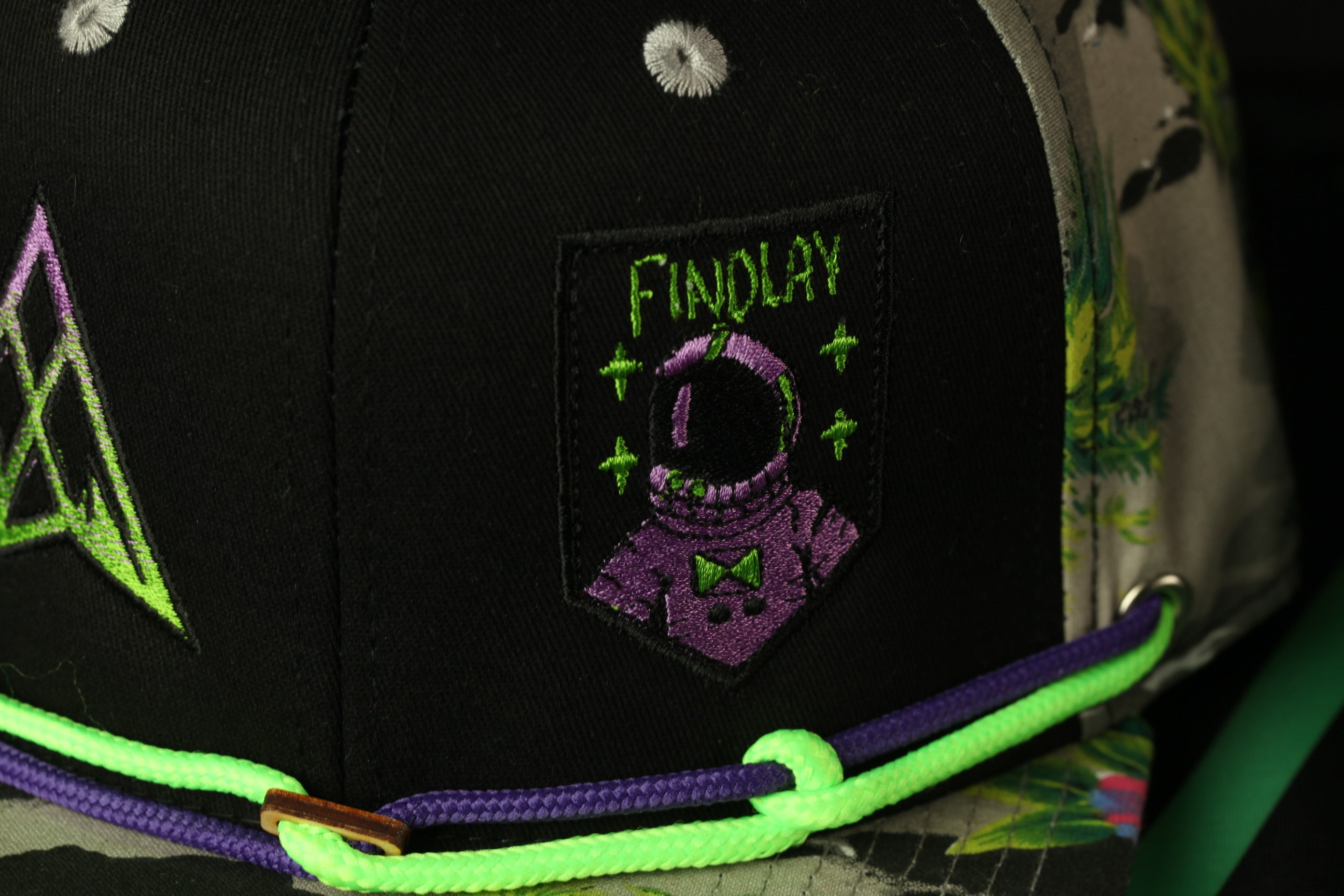 a close-up view of a black baseball cap with a purple and green design, including a small patch with a purple astronaut helmet and the word "FINDLAY" written on it.