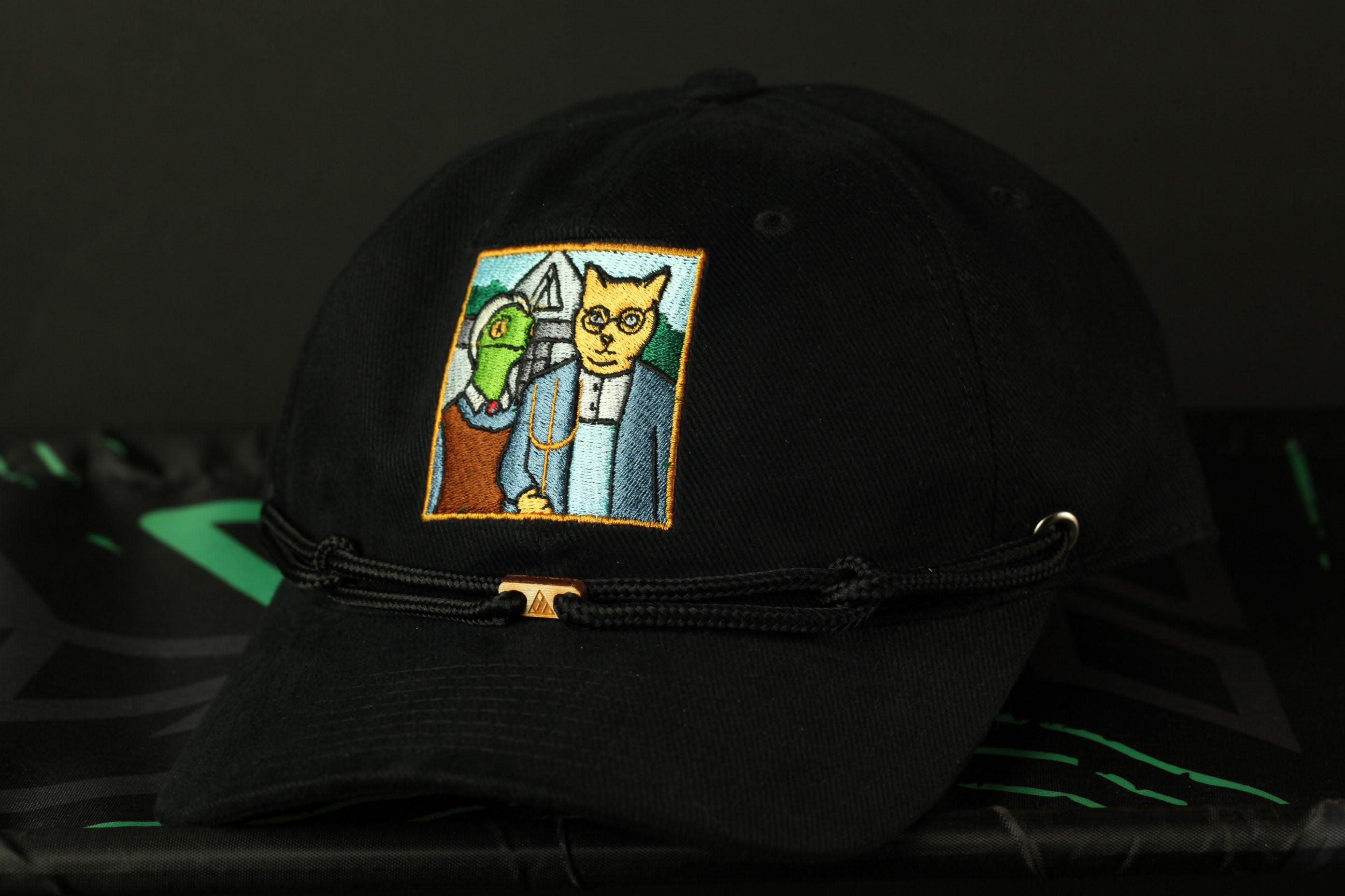 A black baseball cap with a colorful embroidered patch depicting a cat and a fish.