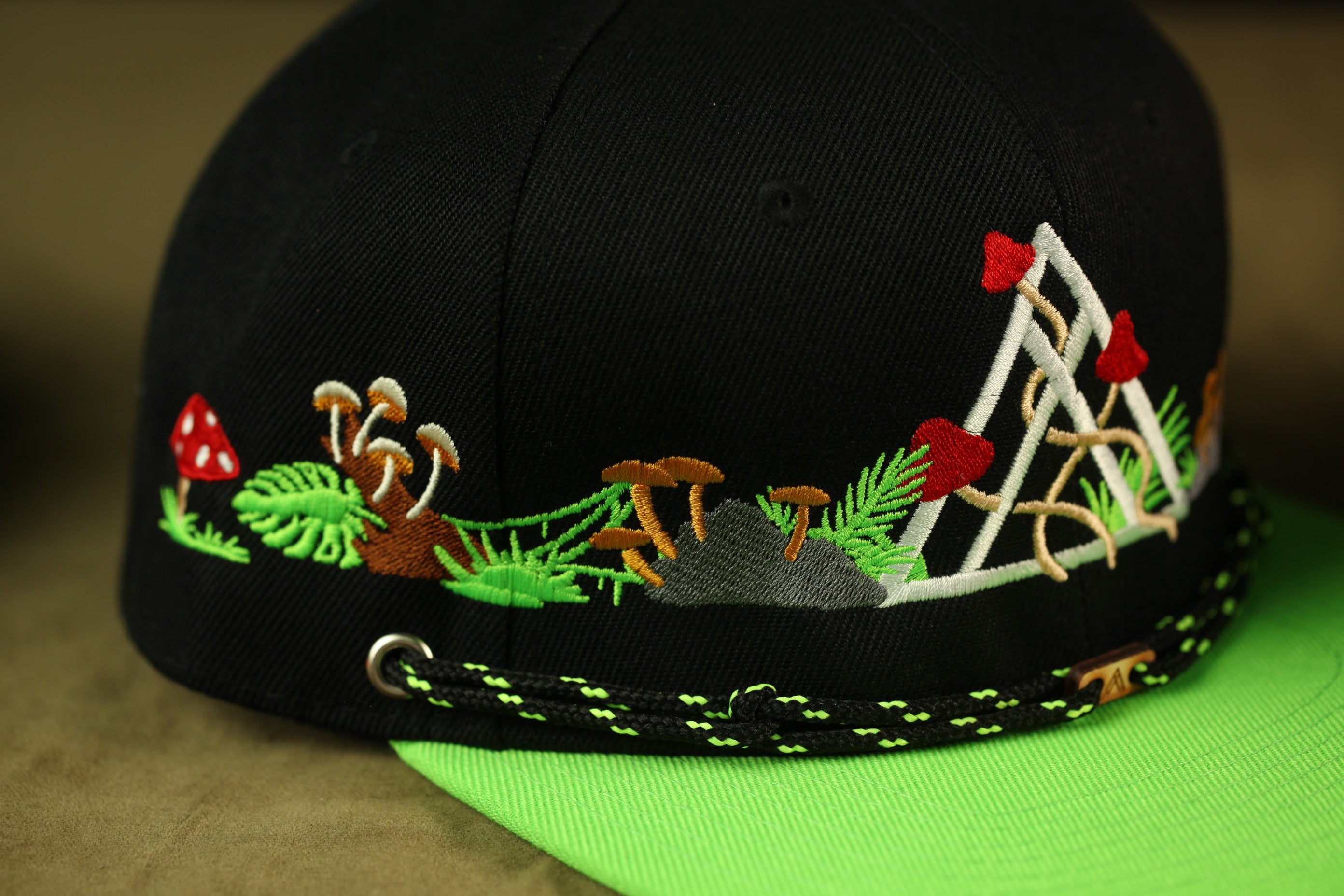 A close-up view of a black baseball cap with a green brim and a decorative pattern of mushrooms, leaves, and other natural elements.