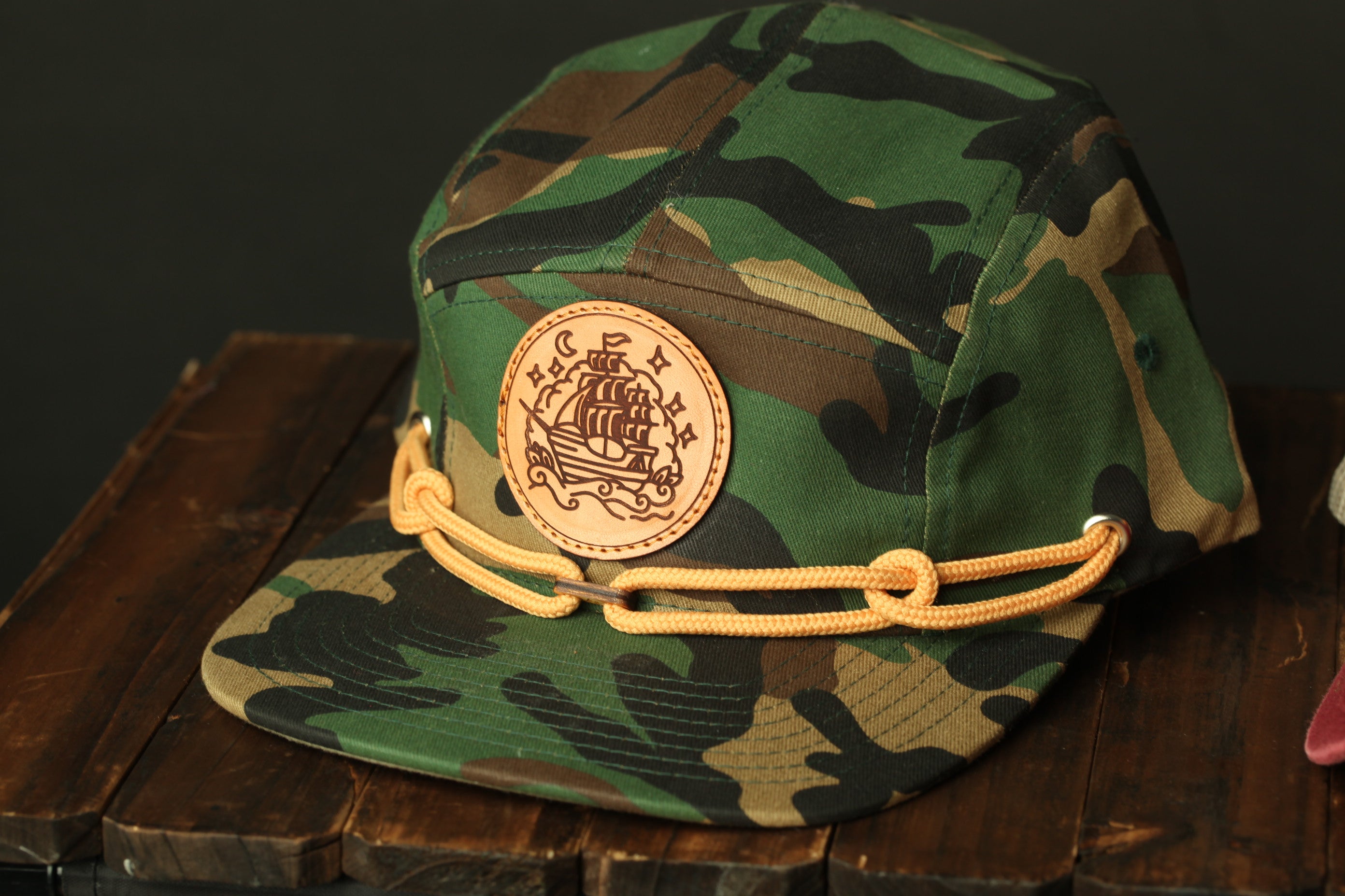 A camouflage baseball cap with a circular patch featuring a ship design and a chain strap.