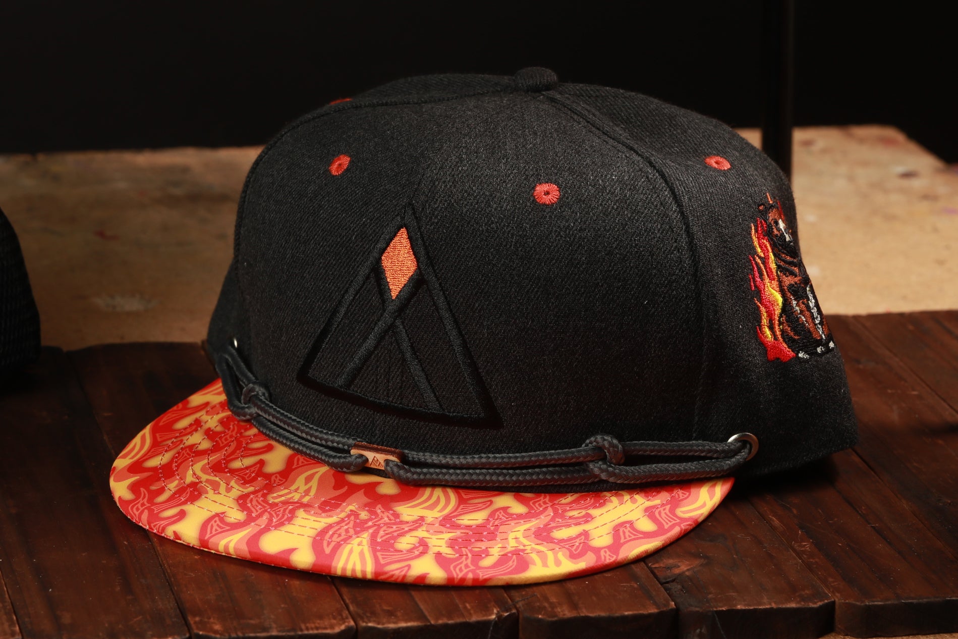A black baseball cap with a red and yellow patterned brim, featuring a triangle design on the front, is displayed on a wooden surface.