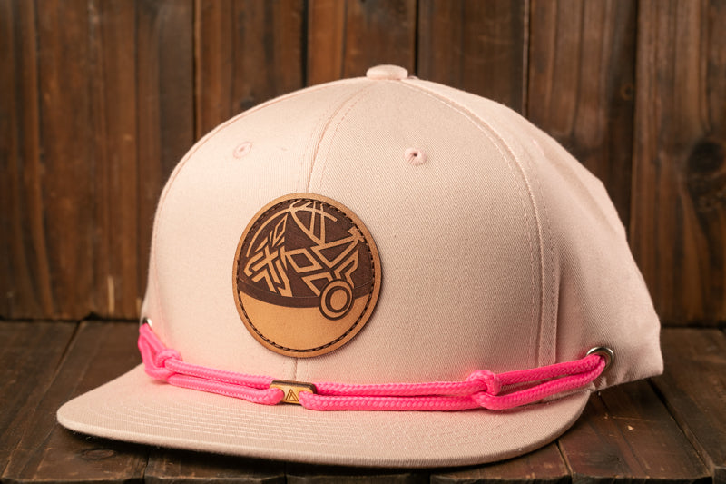 A pink baseball cap with a leather patch and pink braided straps is displayed against a wooden background.