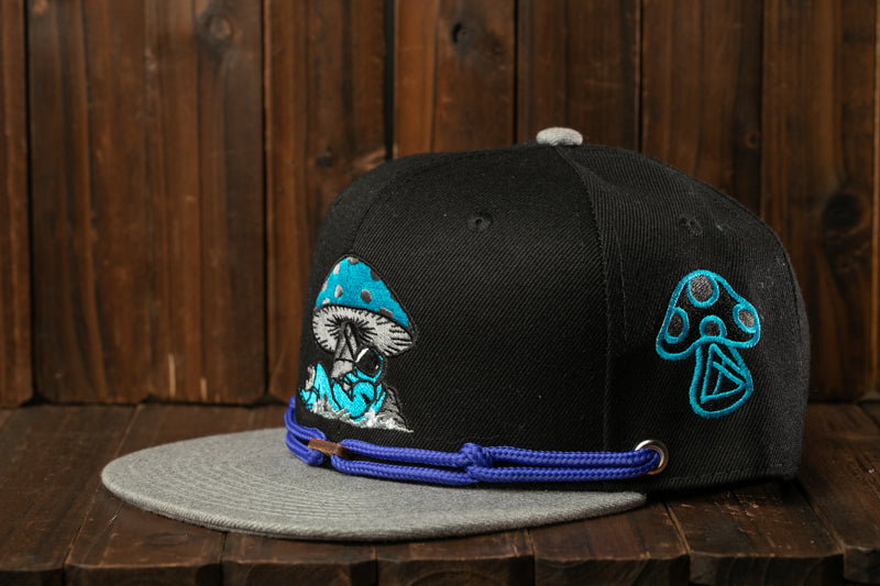 A black baseball cap with blue and teal embroidered designs, including a mushroom and a teardrop, is displayed on a wooden surface.
