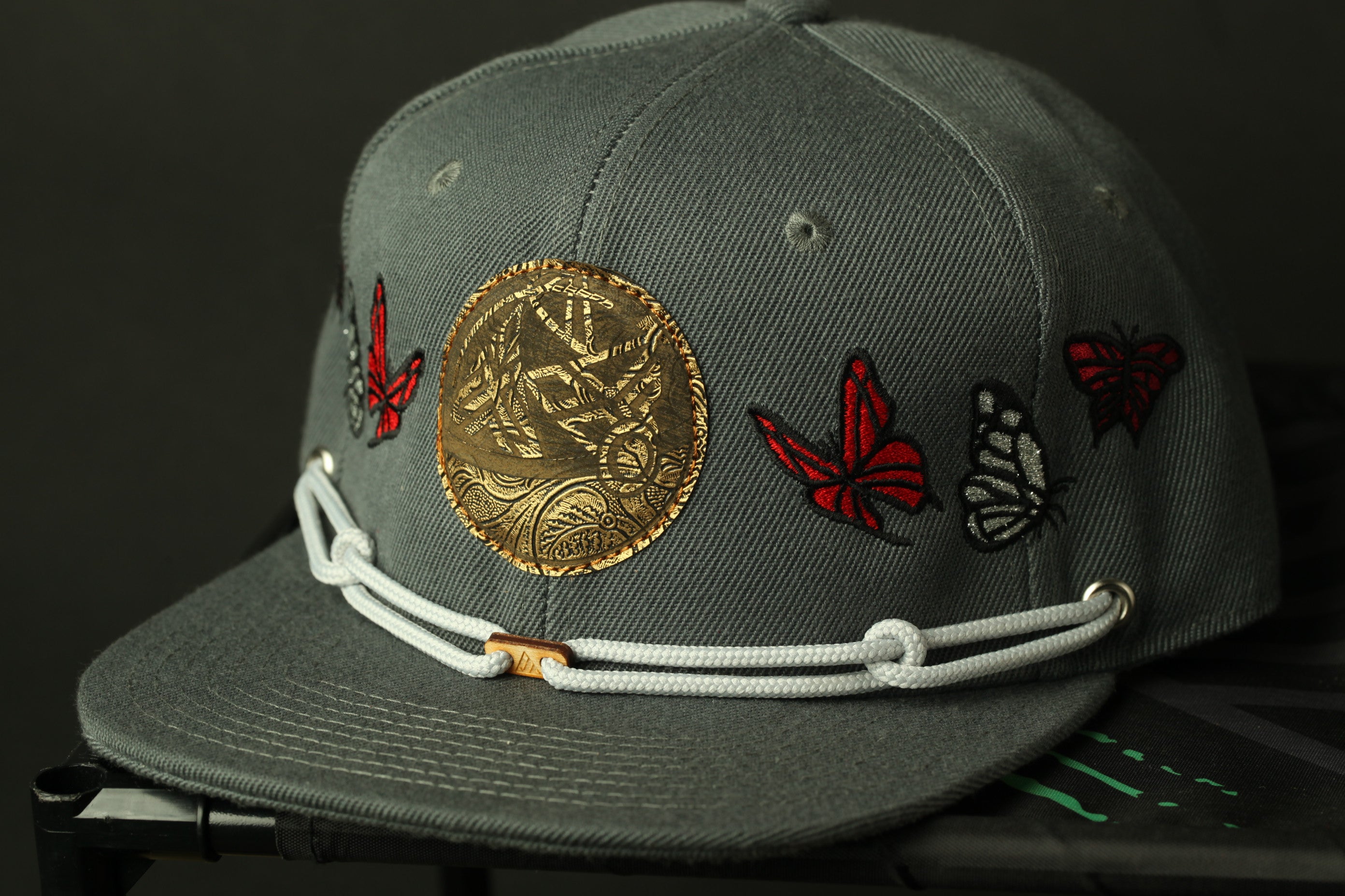 A gray baseball cap with a gold-colored circular patch and red and black butterfly embroidery on the brim.
