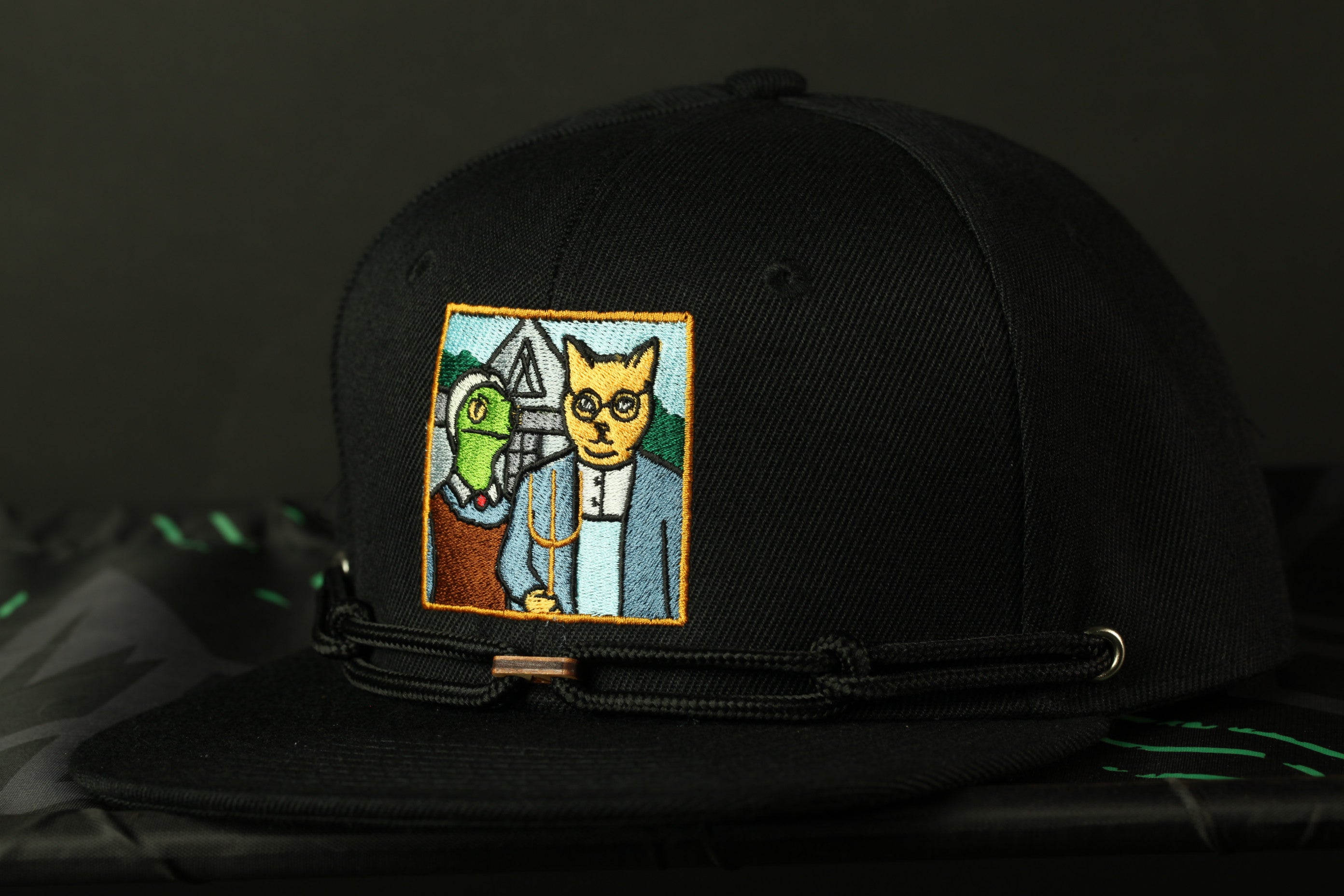 A black baseball cap with a colorful patch featuring a cartoon cat and a green alien.