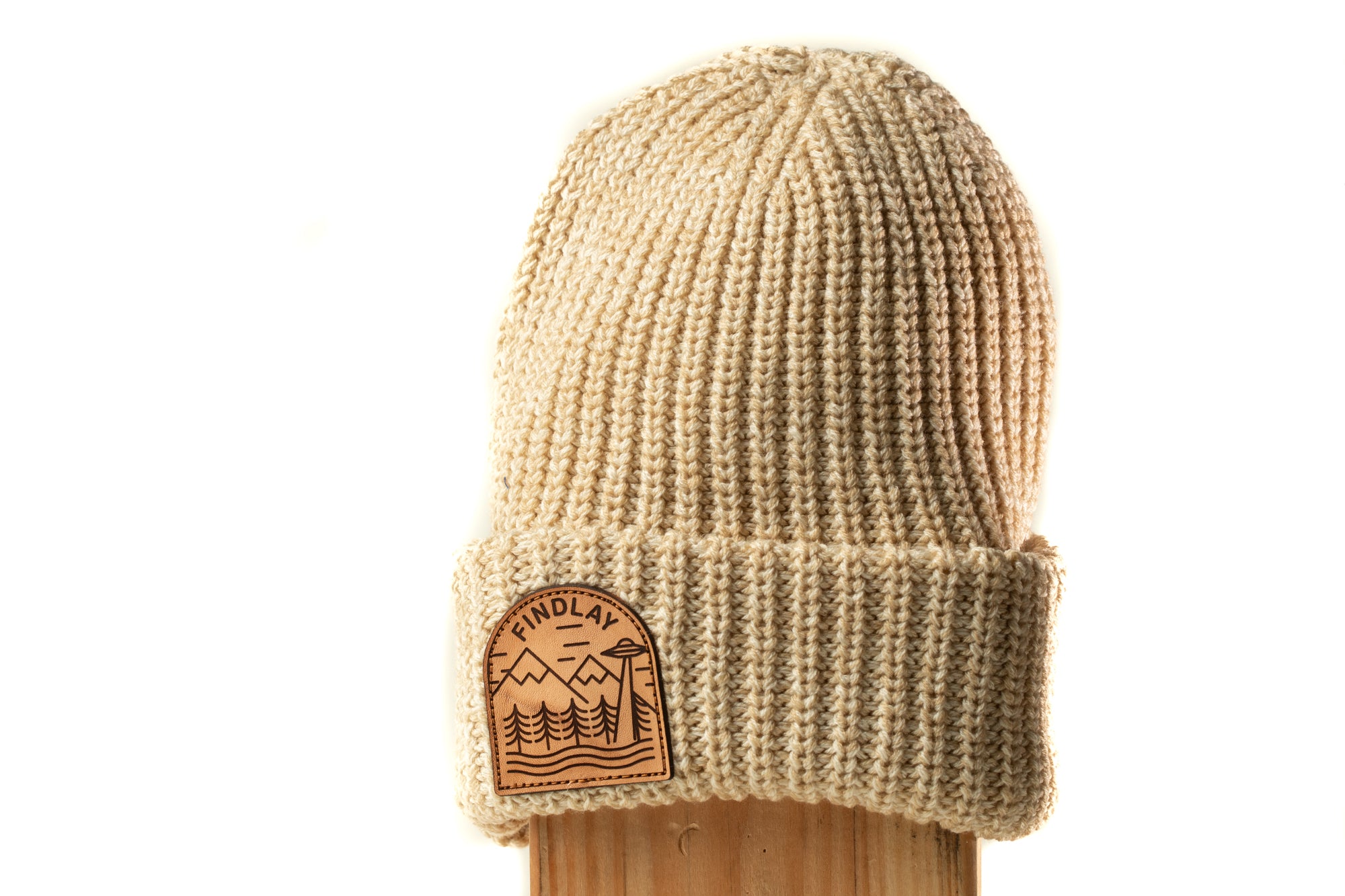 A beige knit beanie with a patch on the front displaying a mountain landscape.