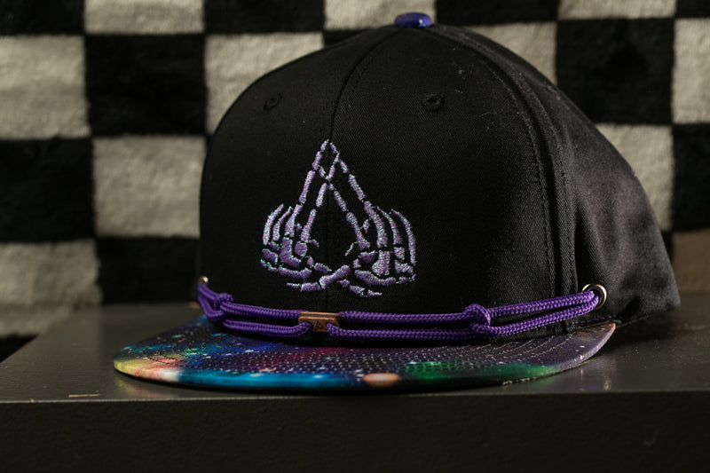 A black baseball cap with a purple band and a colorful, abstract pattern on the brim, placed on a black surface against a checkered background.