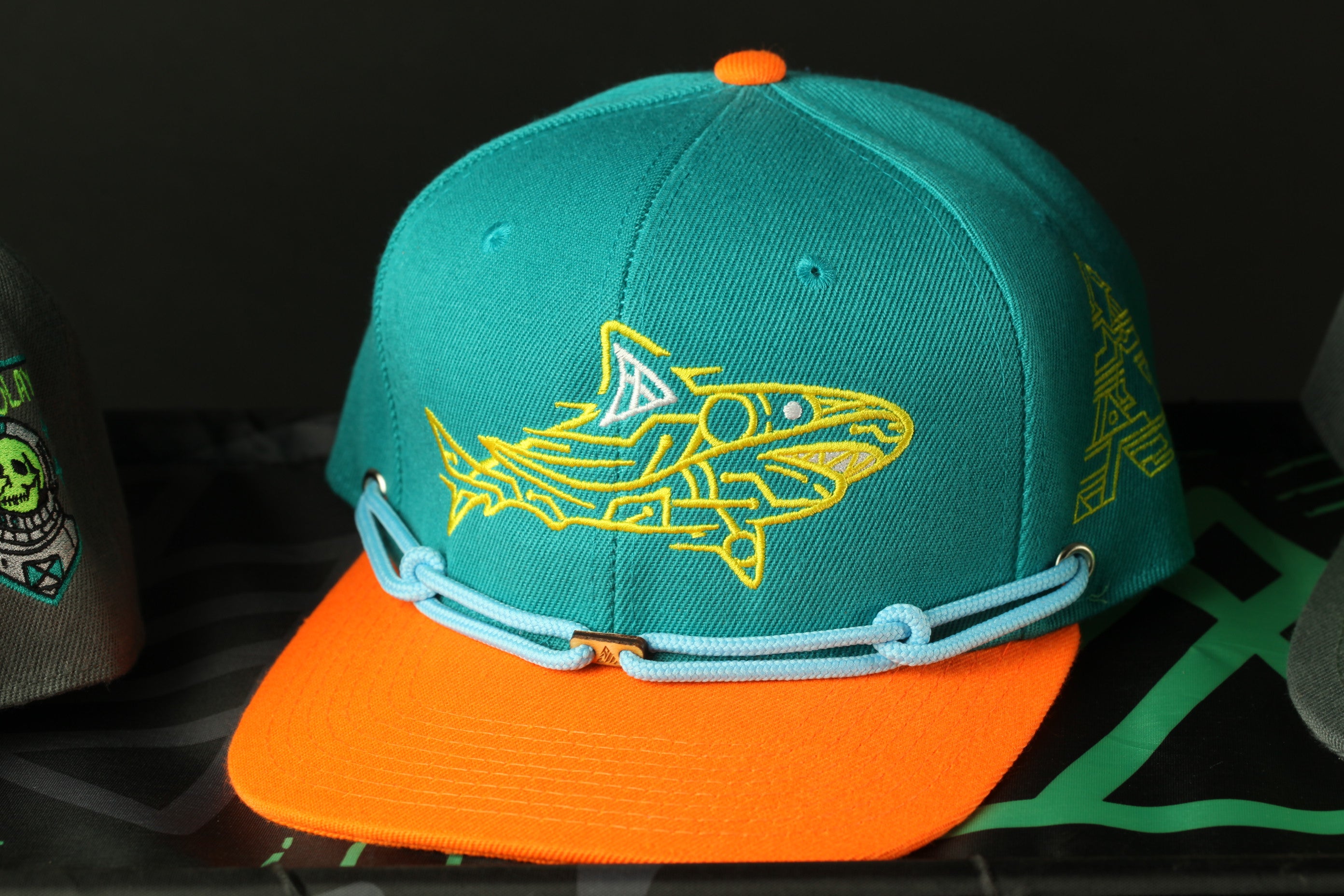 A turquoise baseball cap with a yellow shark design on it, placed on a black surface.