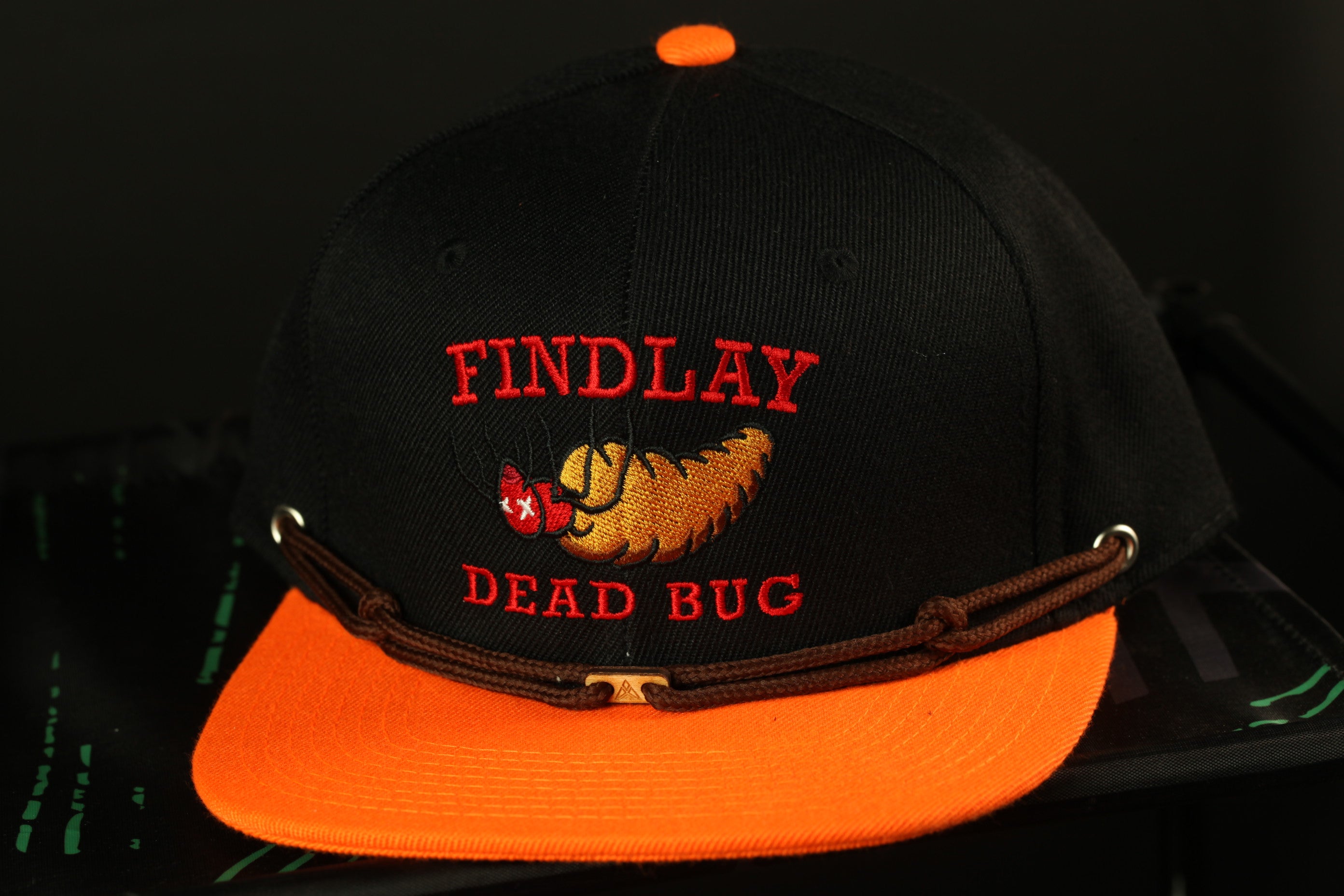 A black baseball cap with an orange brim, featuring a red and yellow design and the text "FINDLAY DEAD BUG" printed on it.