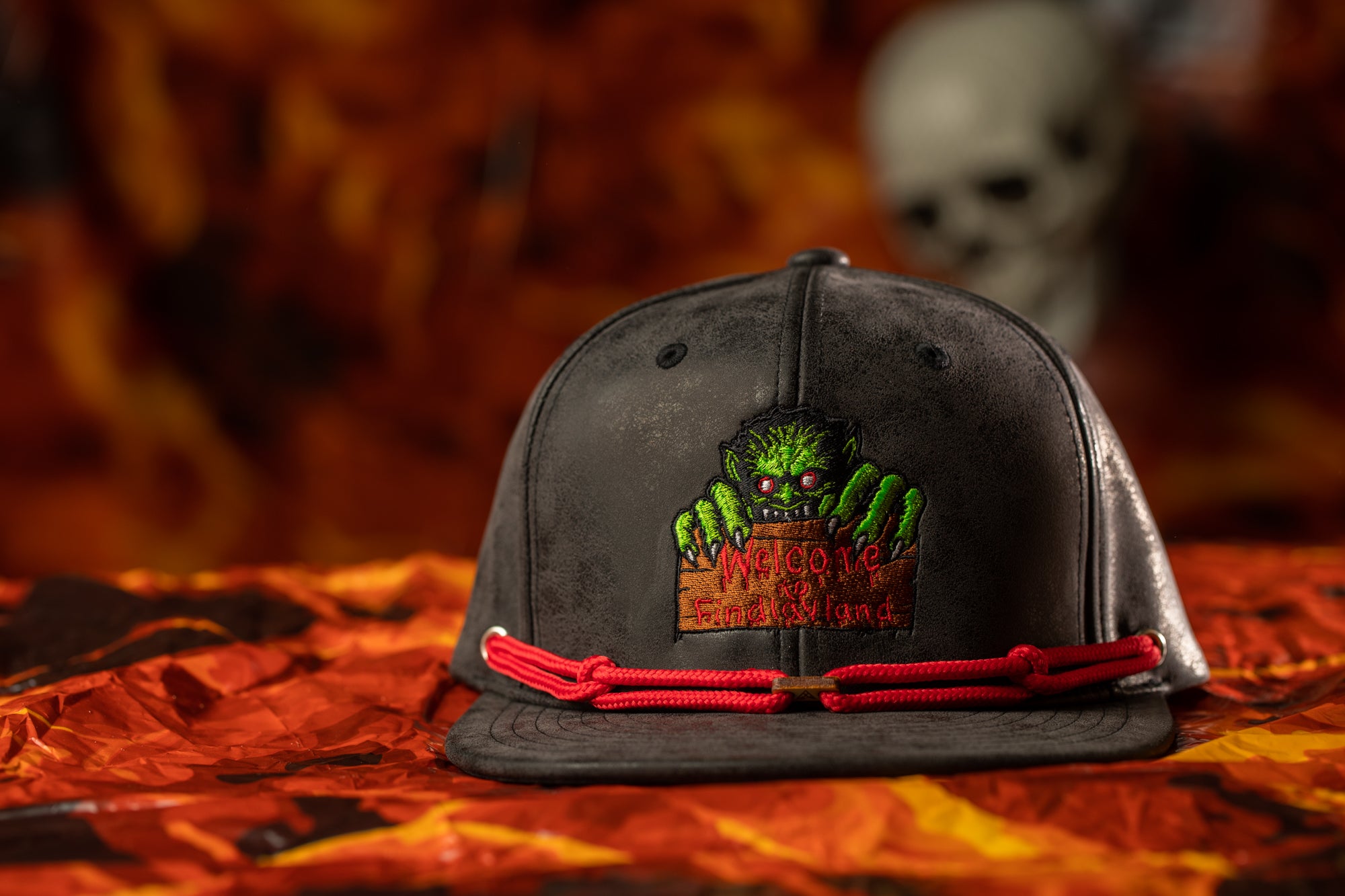 A black baseball cap with a green monster design and a red string around the brim, placed on a red and orange patterned surface.