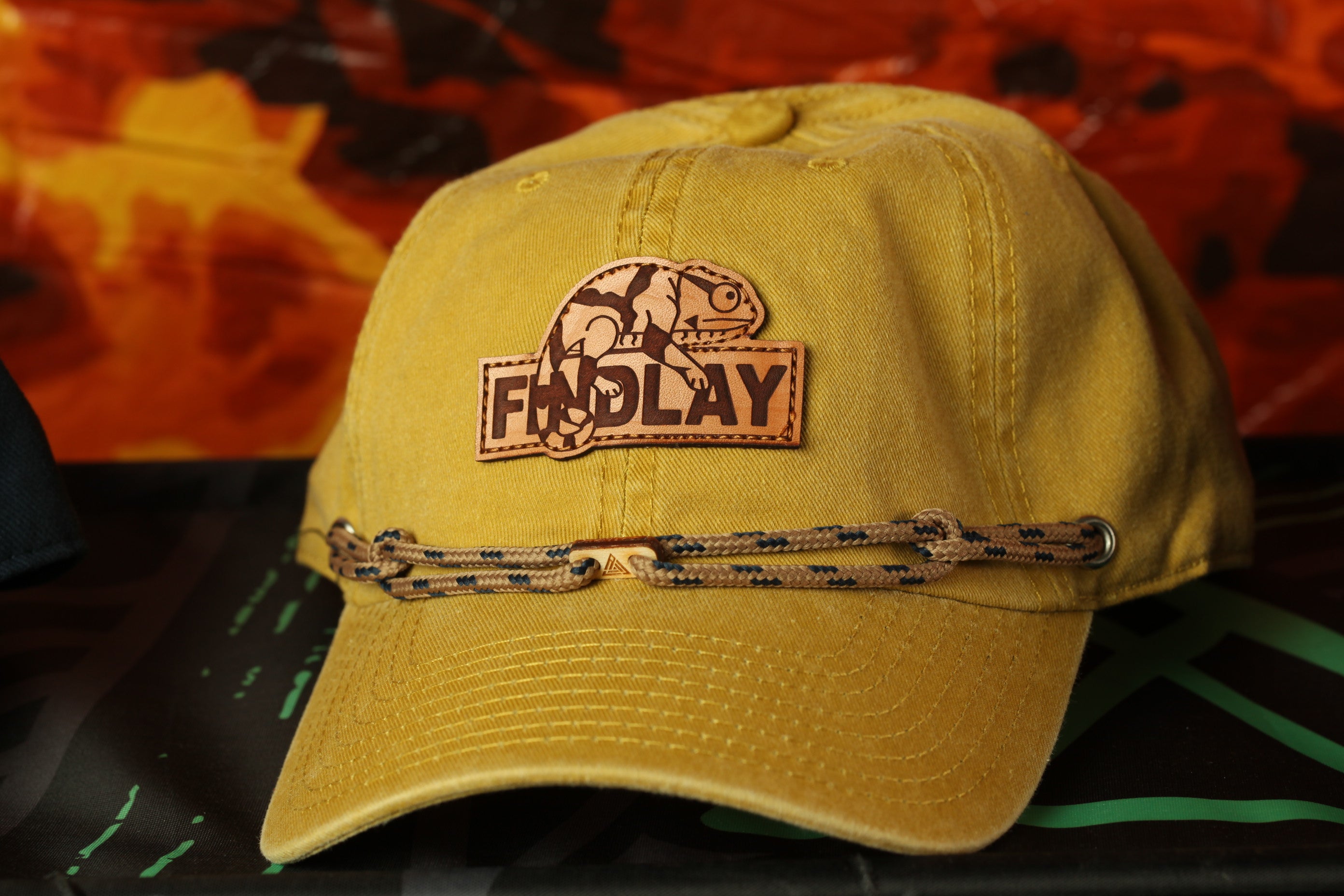 A yellow baseball cap with a leather patch and a black braided strap, featuring the word "FIND LAY" on the front.