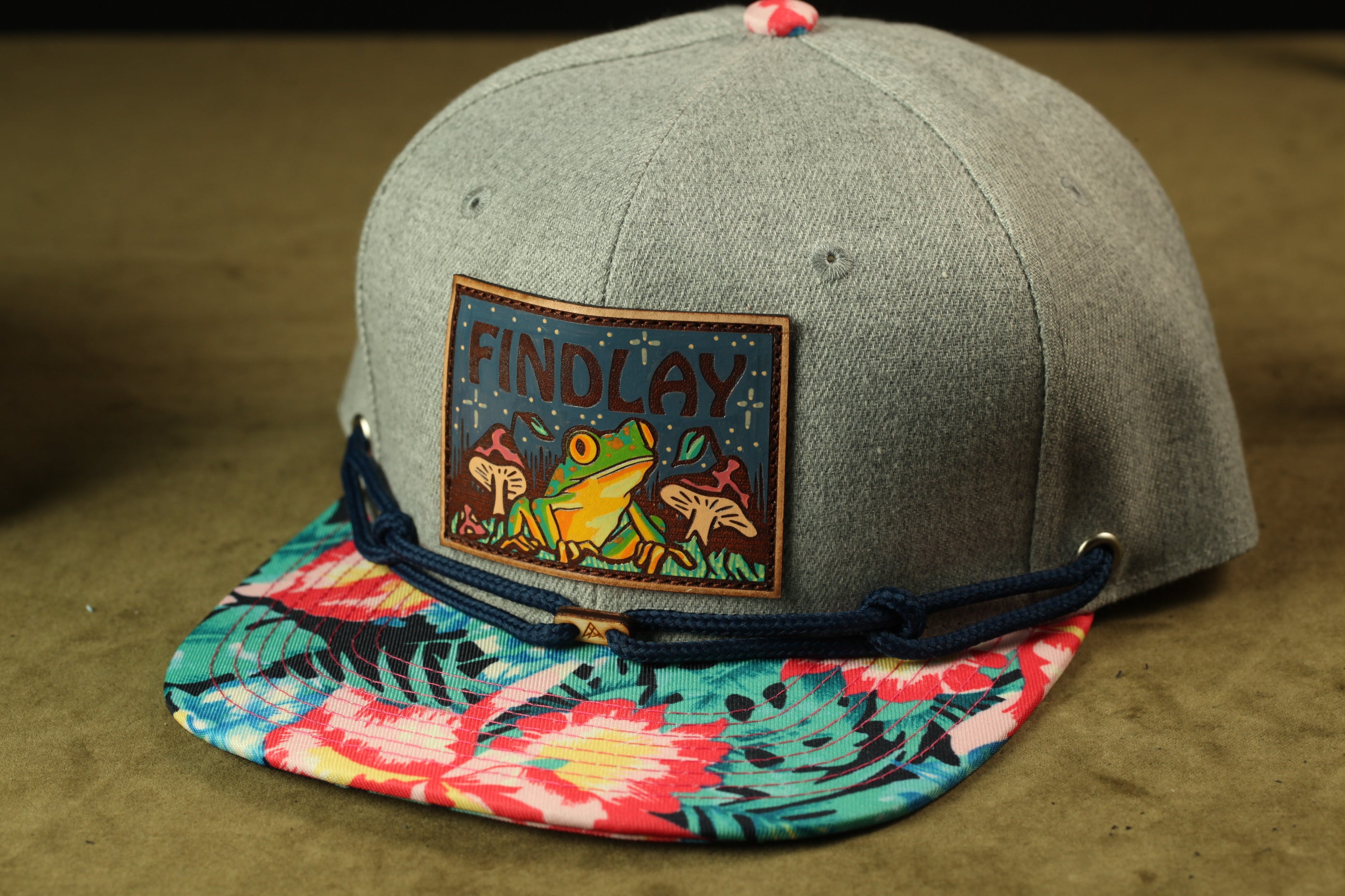 A gray baseball cap with a colorful floral pattern and a patch featuring a frog and mushrooms.