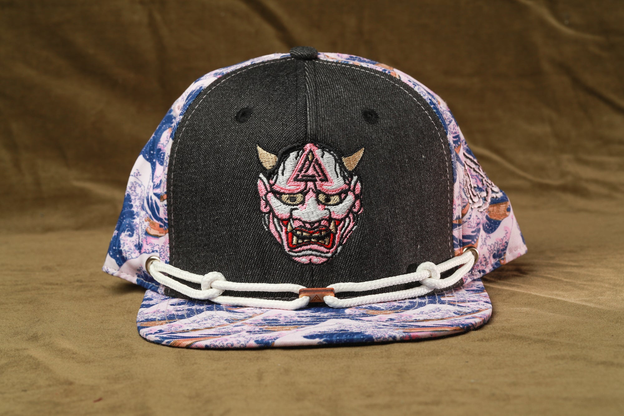 A baseball cap with a black brim and a colorful, patterned fabric on the front. The cap has a white chain strap and a graphic of a mask or mask on the front.