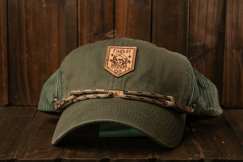 A green baseball cap with a leather patch on the front, sitting on a wooden surface.