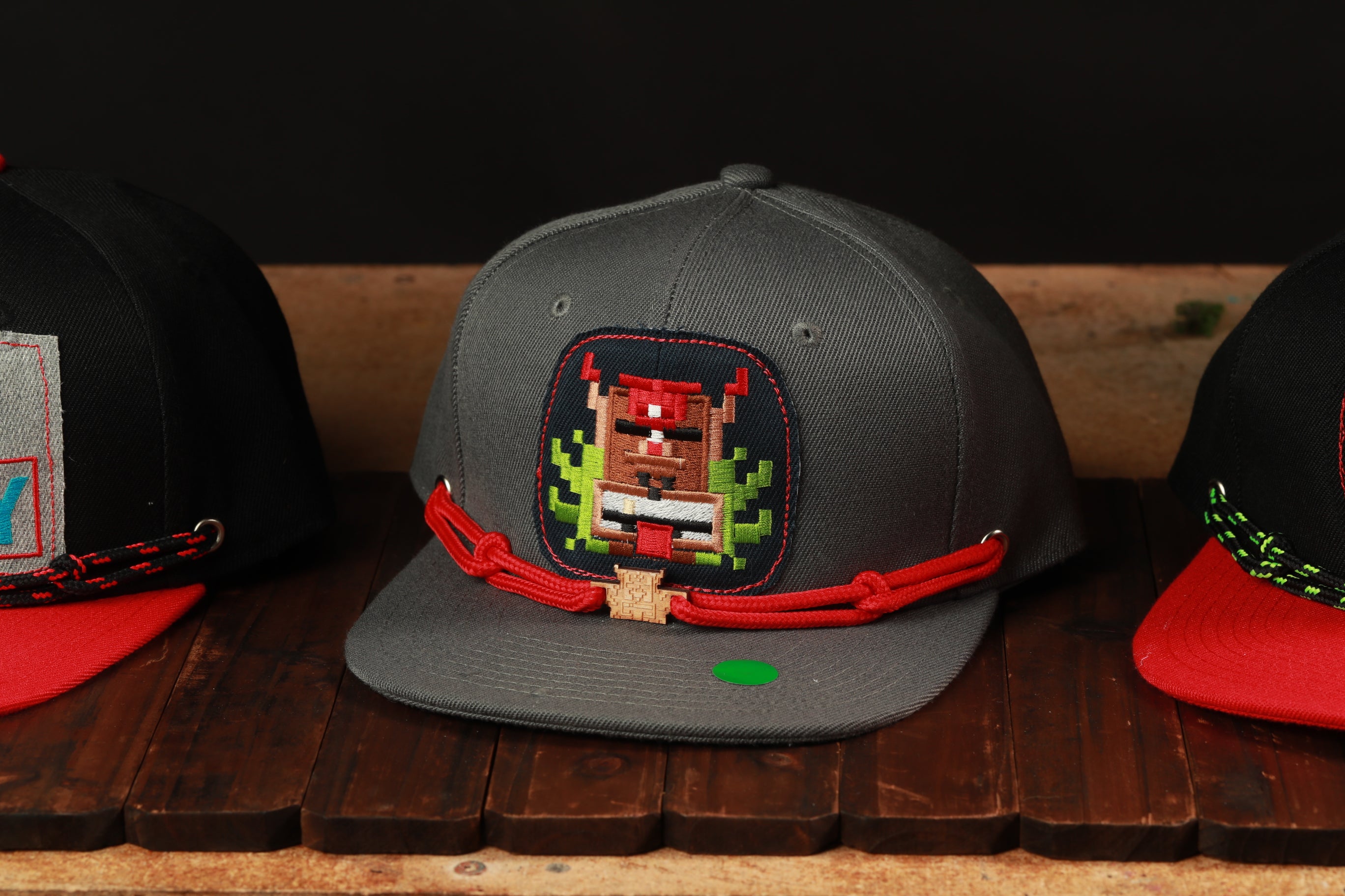 A gray baseball cap with a red and green pixelated character design on the front, displayed on a wooden surface.