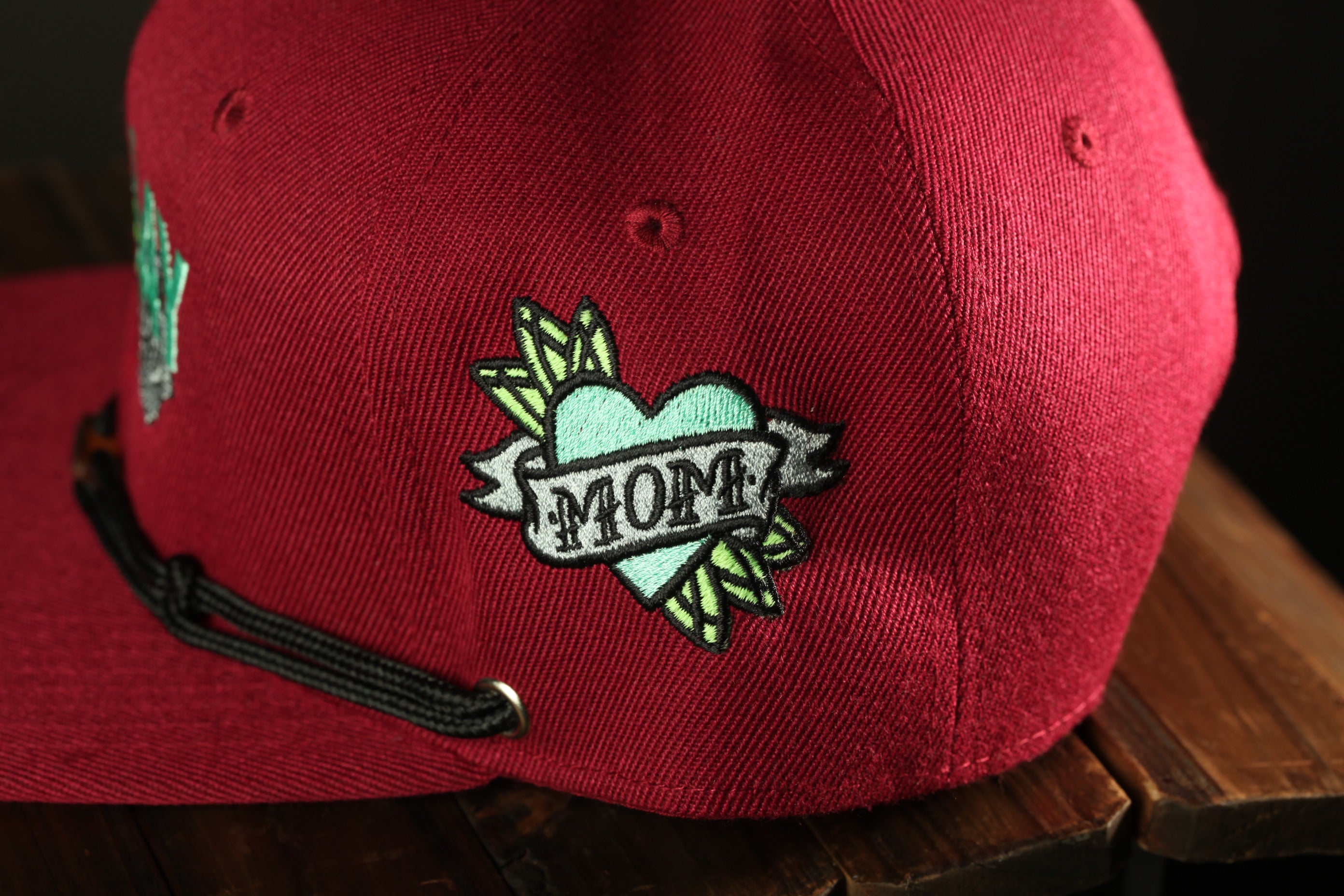 A close-up of a red baseball cap with a heart-shaped embroidered patch that says "MOM" on it.