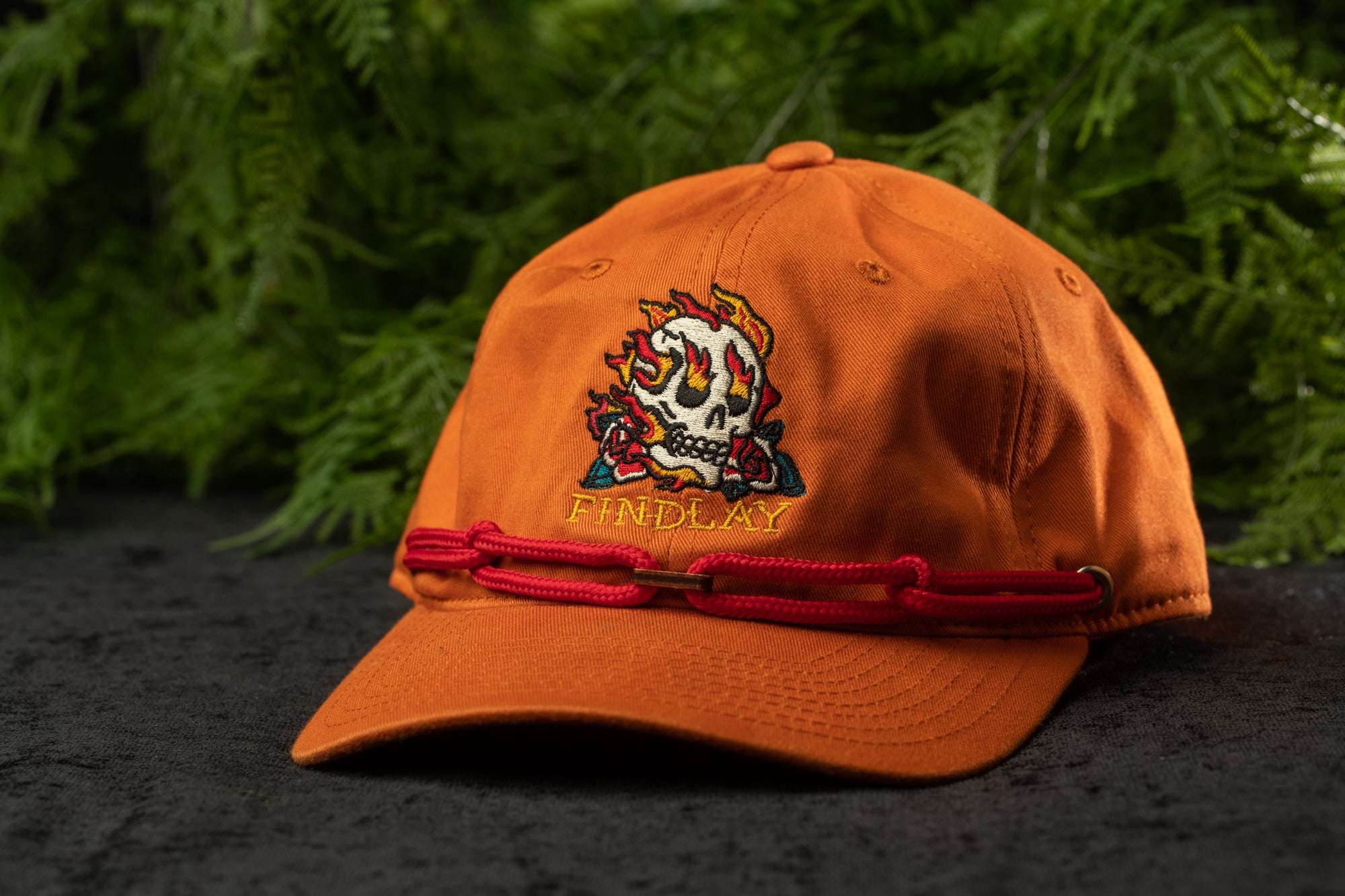 An orange baseball cap with a skull design and the word "FINDA" printed on it, placed on a surface with green foliage in the background.
