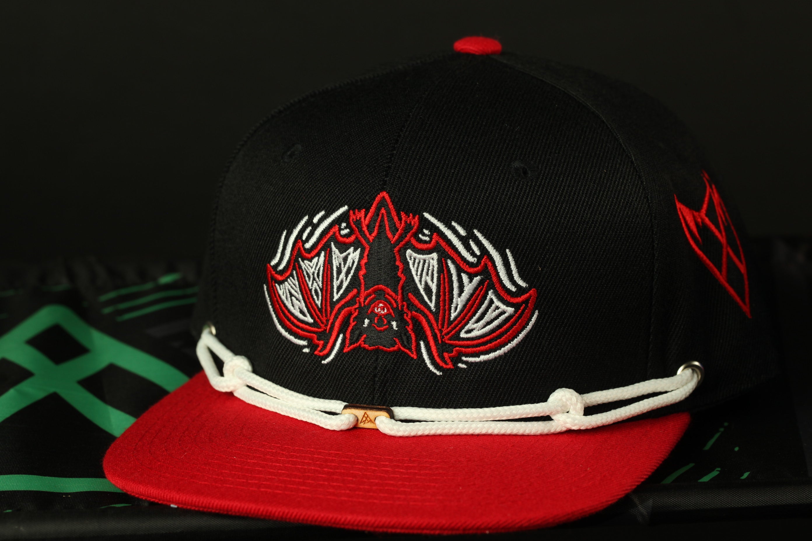 A black baseball cap with a red brim and a bat-like design on the front, resting on a black surface.