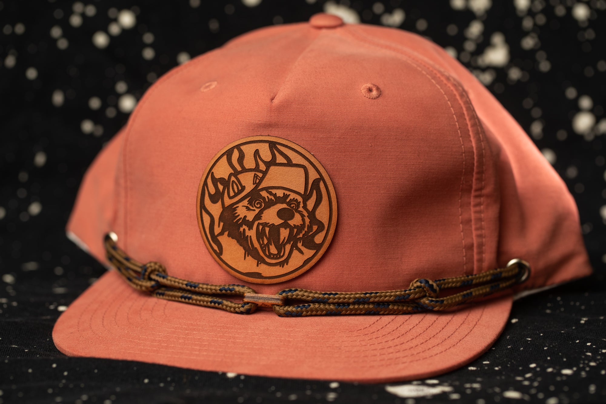 A pink baseball cap with a brown leather patch featuring a cartoon character on it, placed against a black background with white speckles.