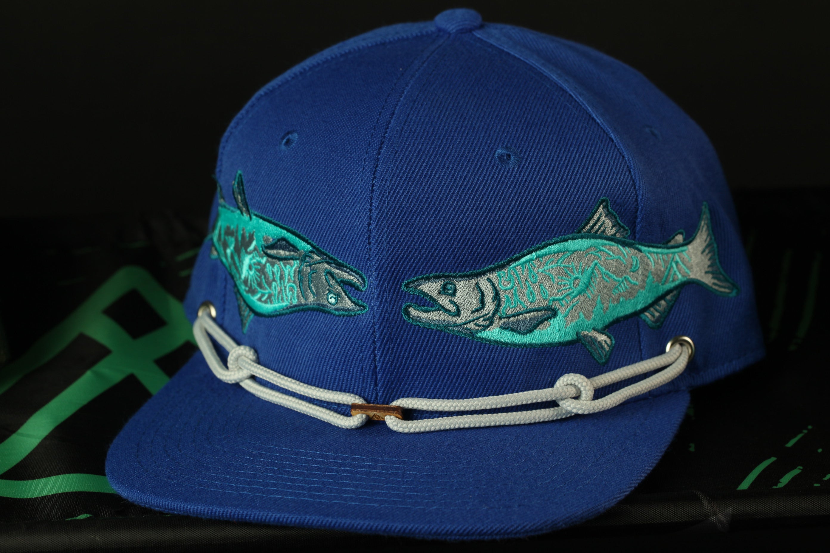 A blue baseball cap with a fish embroidered on it, featuring two fish designs on either side of the brim.