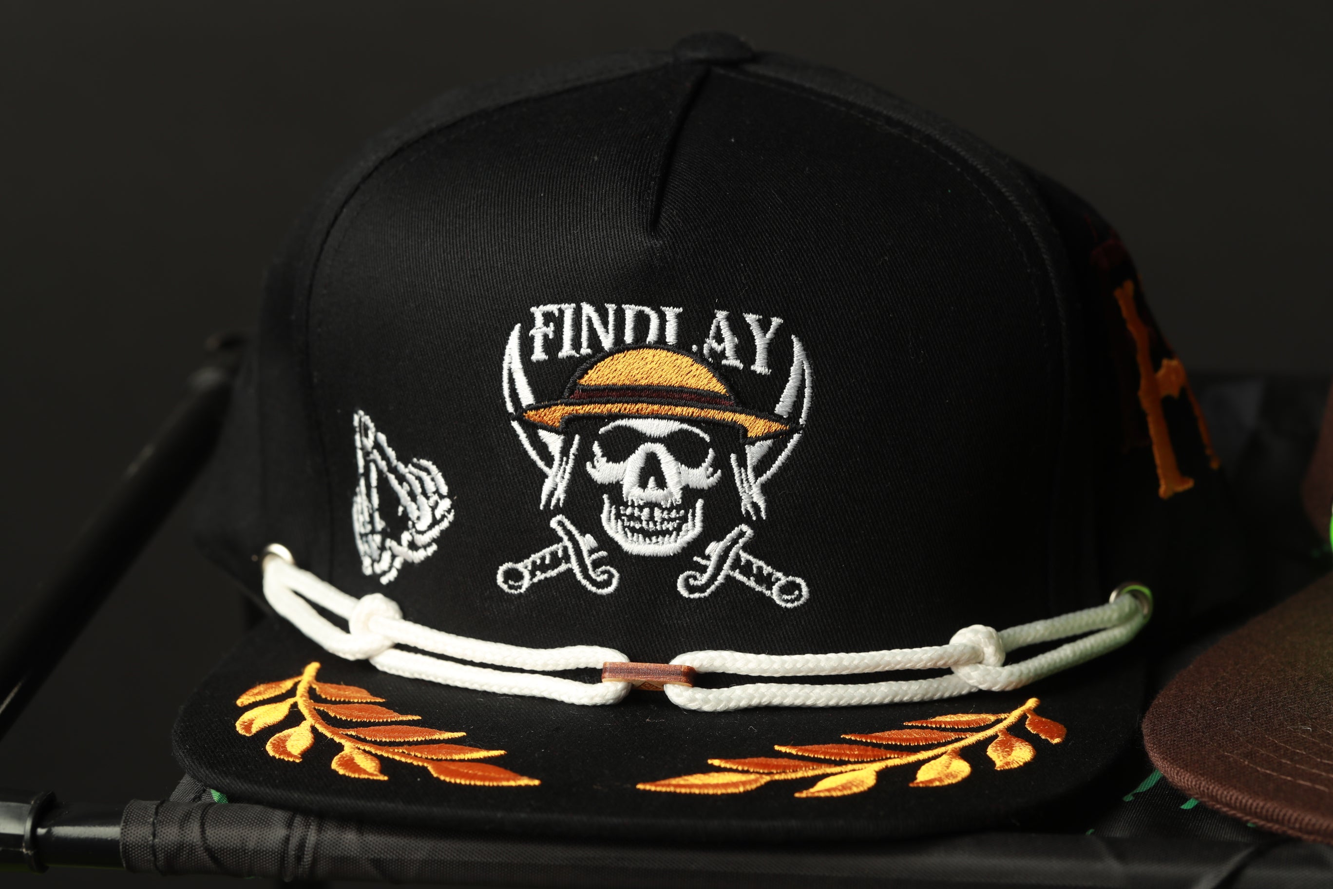 A black baseball cap with a skull and crossbones design, a chain, and a laurel wreath embroidered on it.