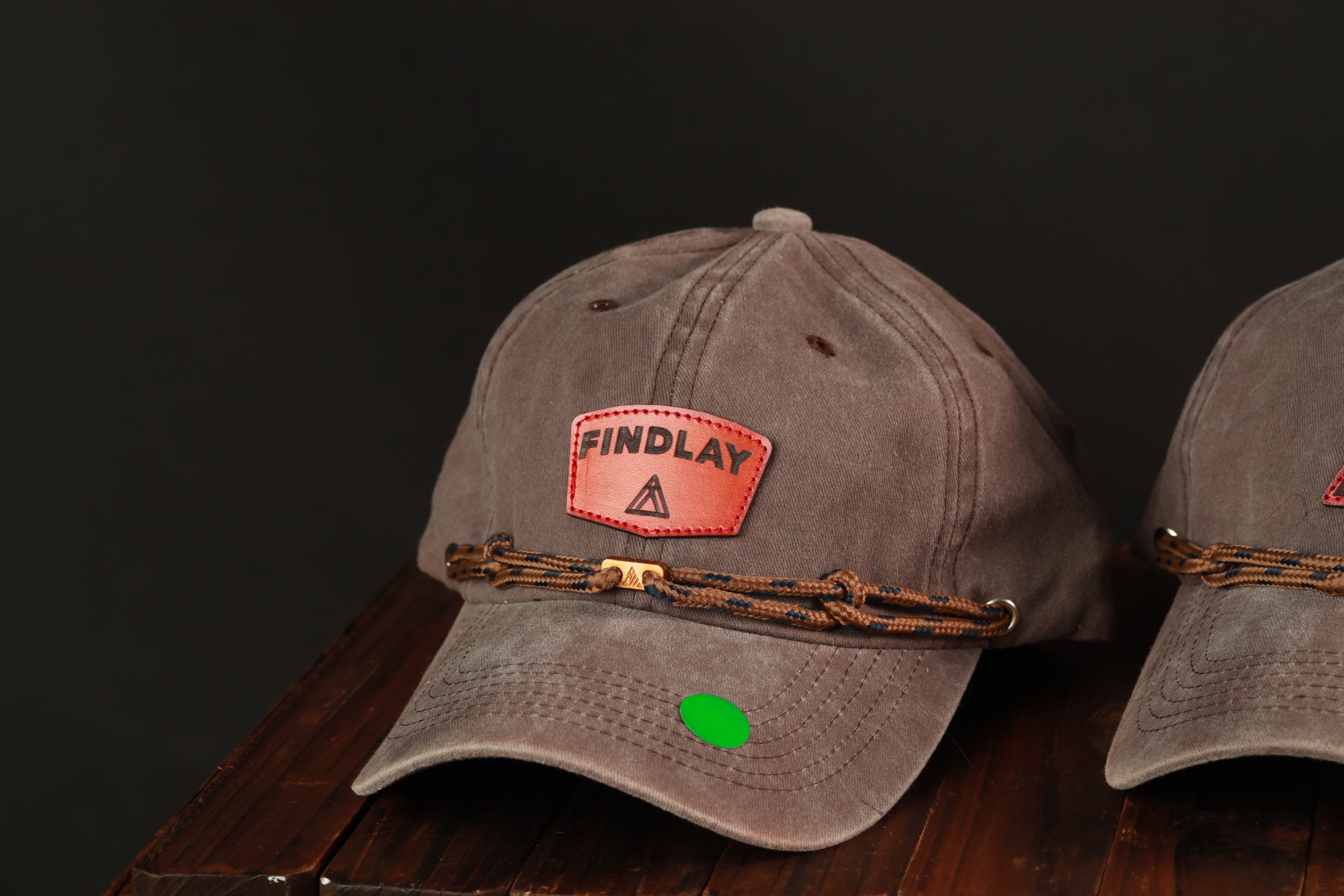 A close-up image of a brown baseball cap with a red "FINDLAY" patch and a green tag on the front.