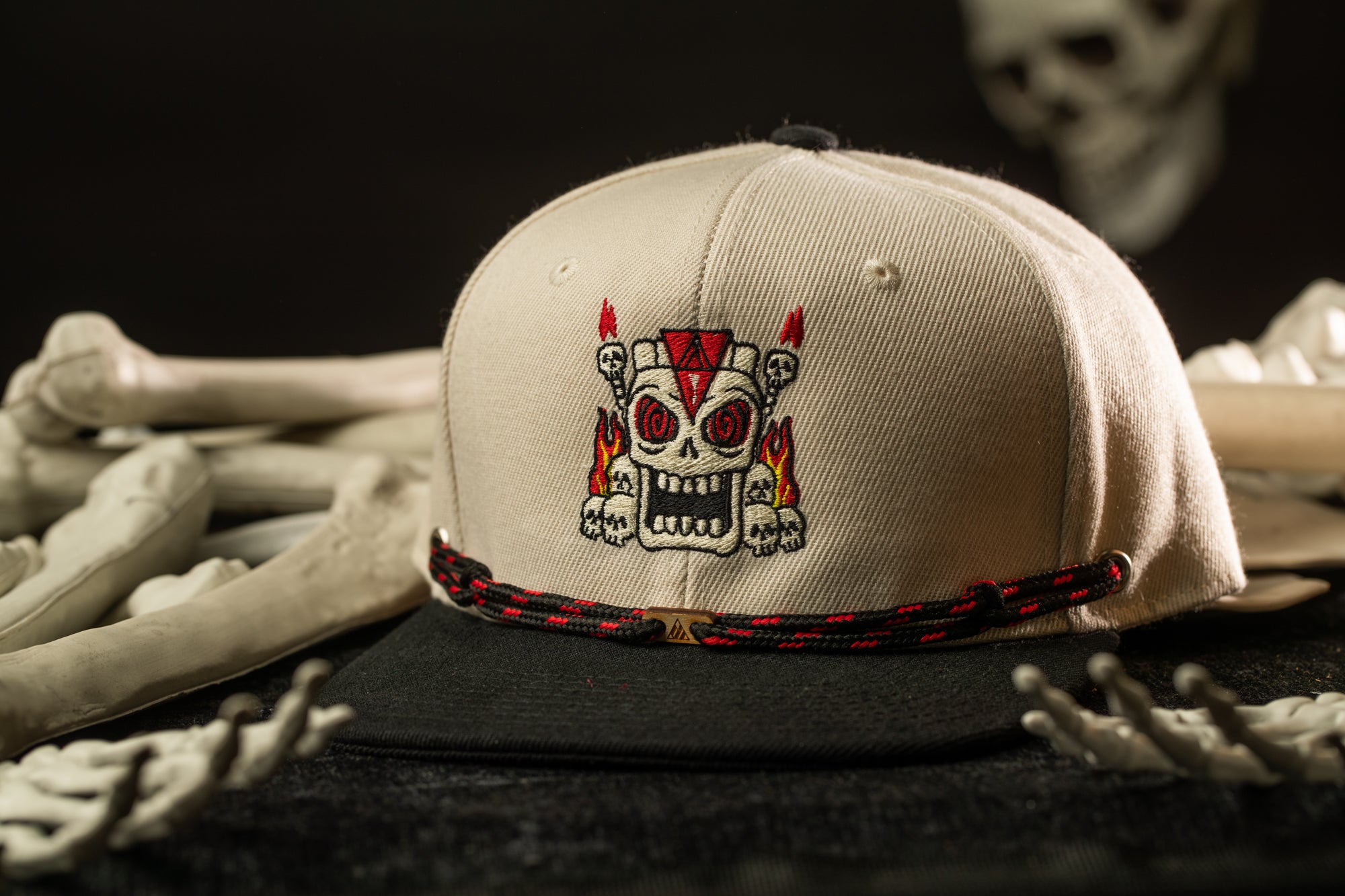A beige baseball cap with a skull design on the front, placed on a surface with various bones and skulls in the background.