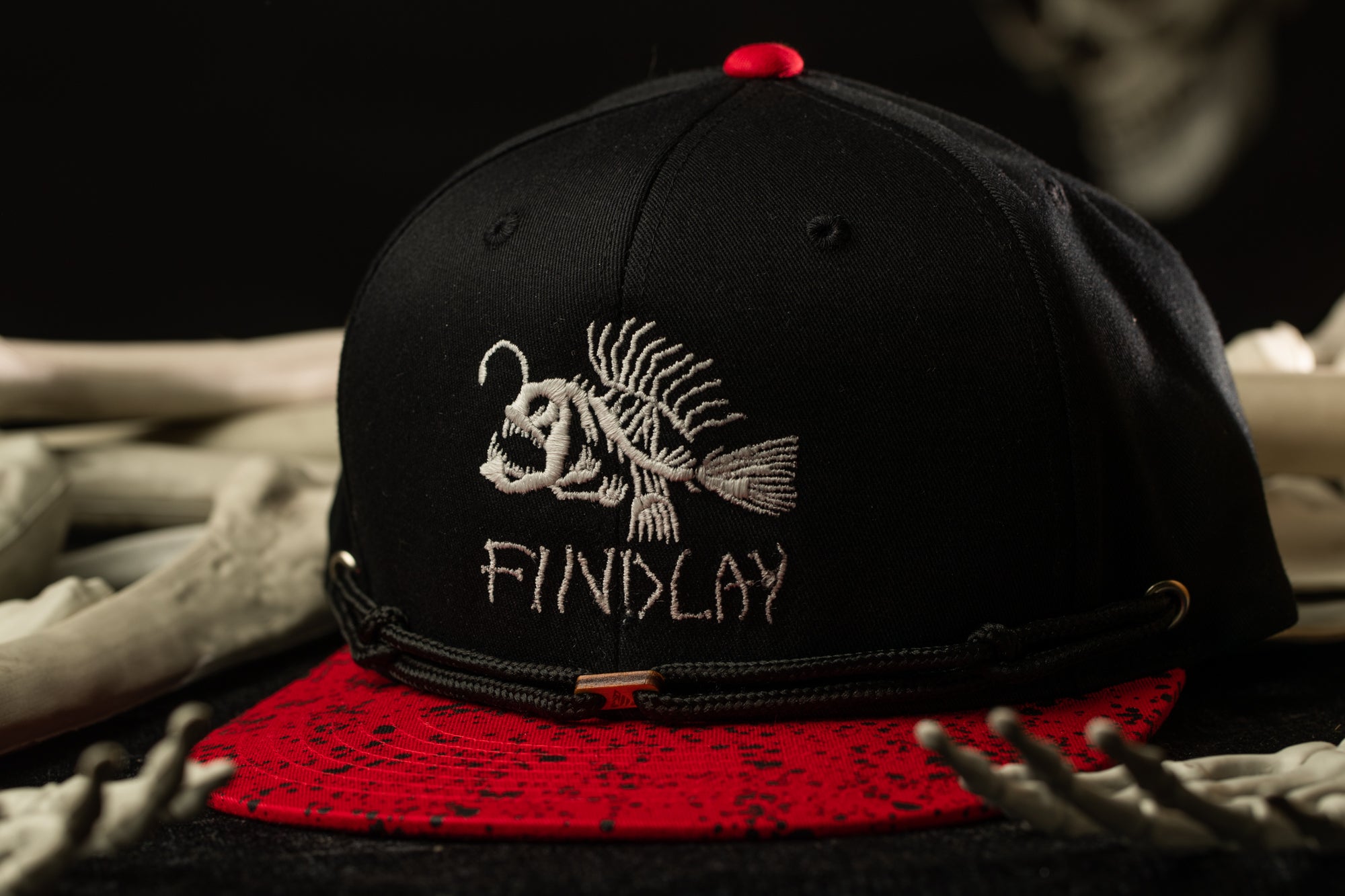 A black baseball cap with a red brim and a white fish embroidered on it, placed on a red and black patterned surface.
