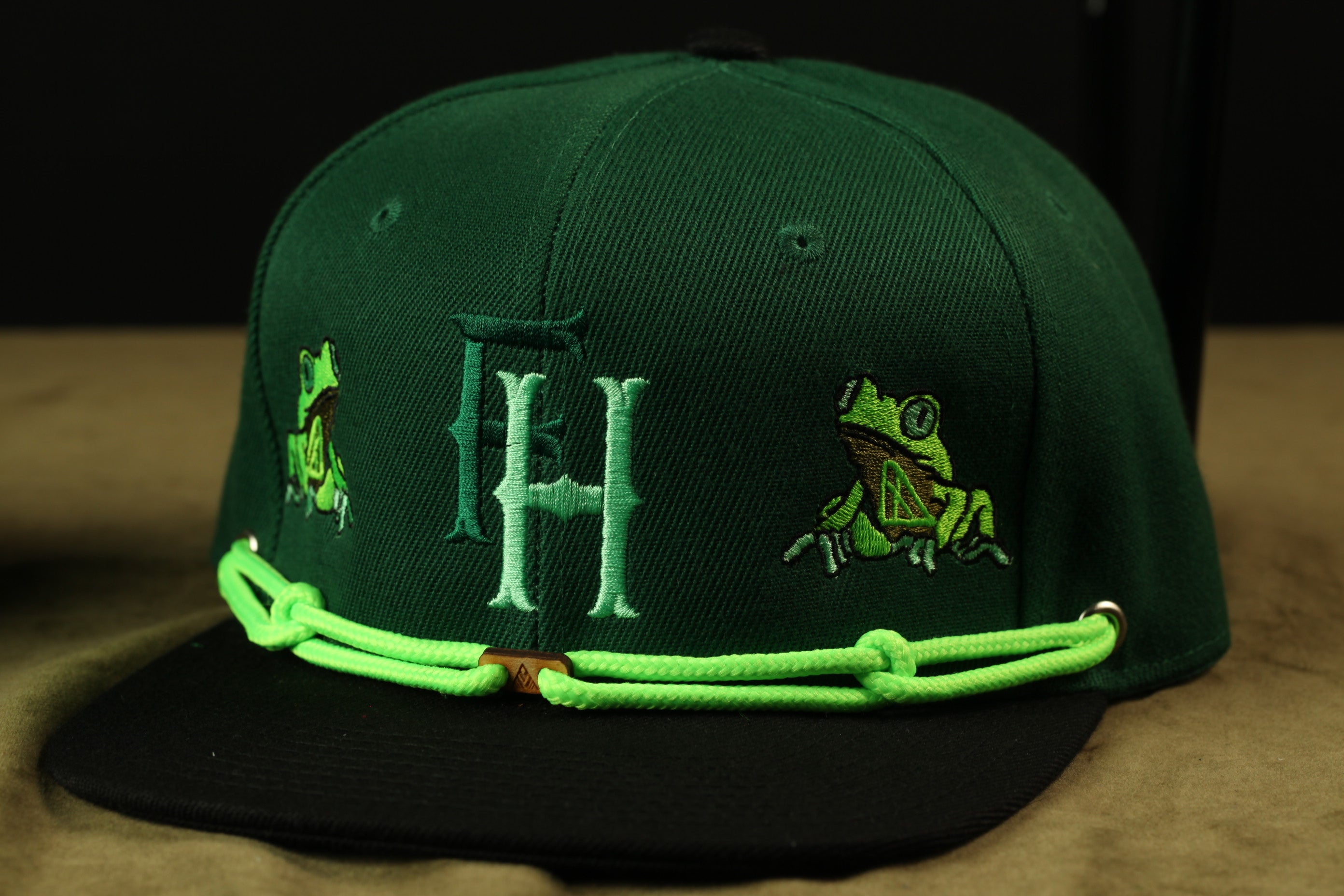 A green baseball cap with a frog embroidered on it, featuring a black brim and a green chain strap.