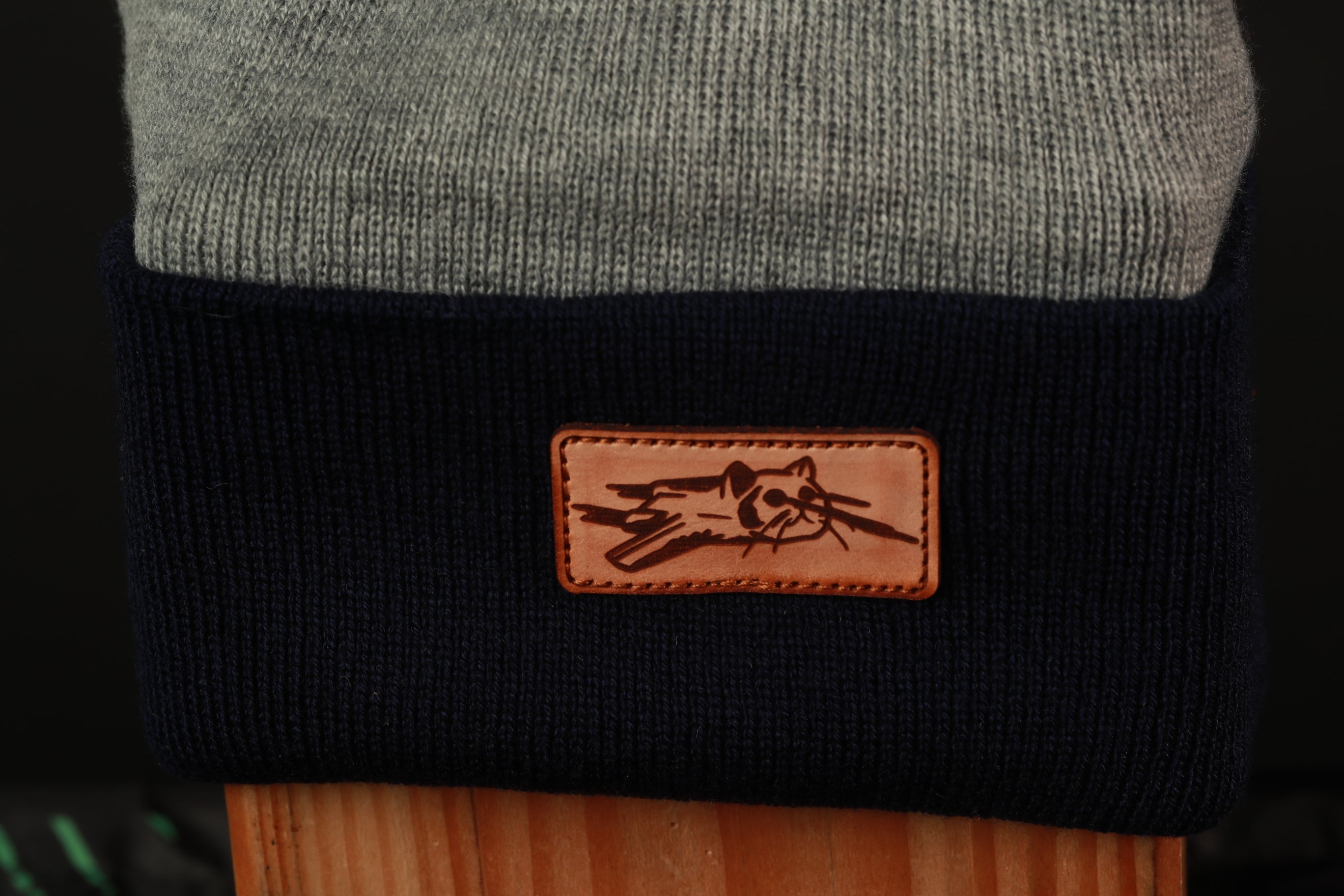 A close-up view of a gray and black knitted or crocheted hat with a leather patch featuring a cat design on it.