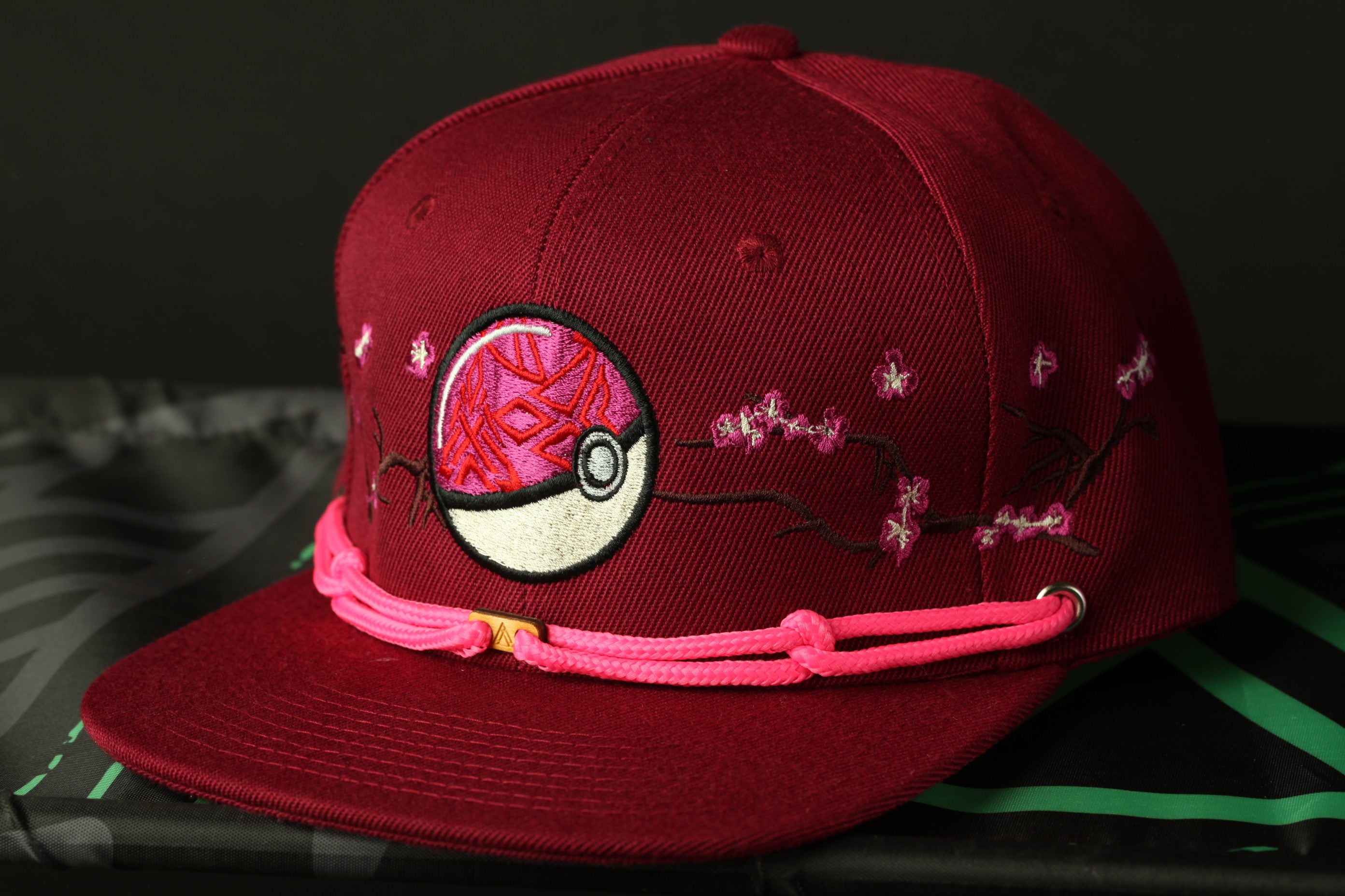 A maroon baseball cap with a pink braided strap and a Pokemon-inspired design on the front.