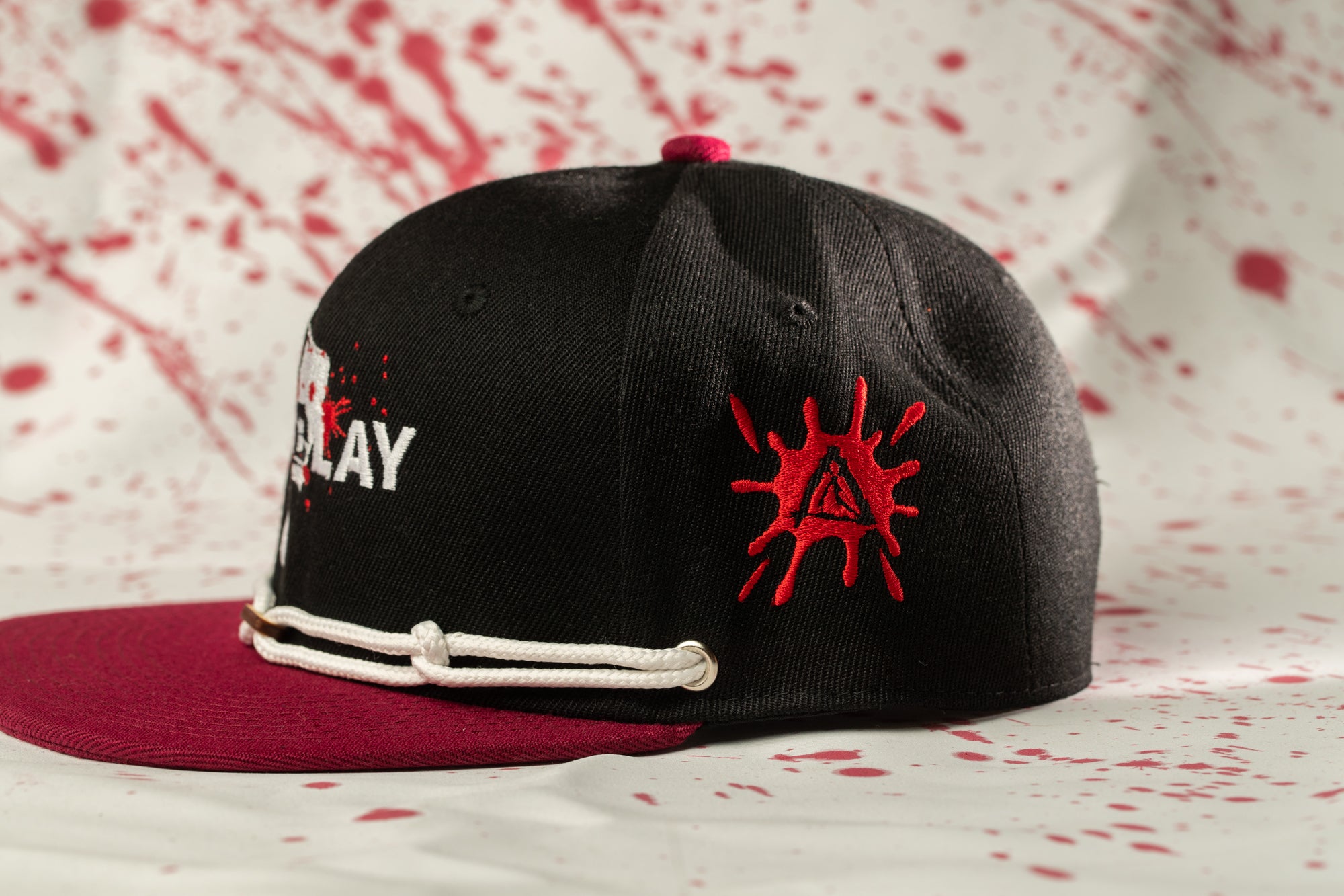 A black baseball cap with a red splash design on the side, placed on a white surface with red splatters.