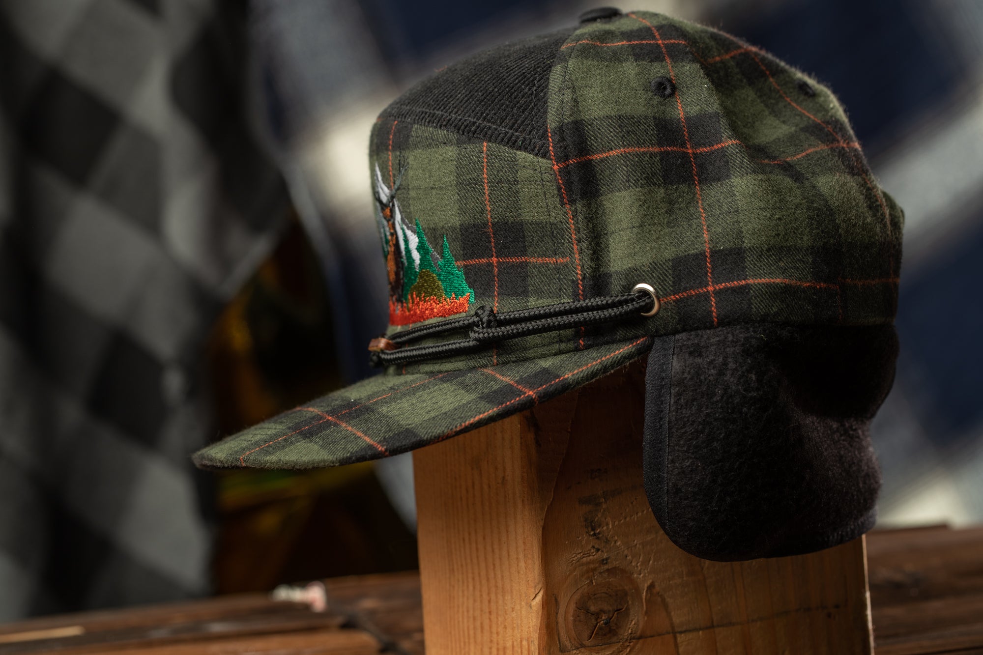 A plaid baseball cap with a black brim and a small embroidered logo on the front, displayed on a wooden stand against a blurred background.