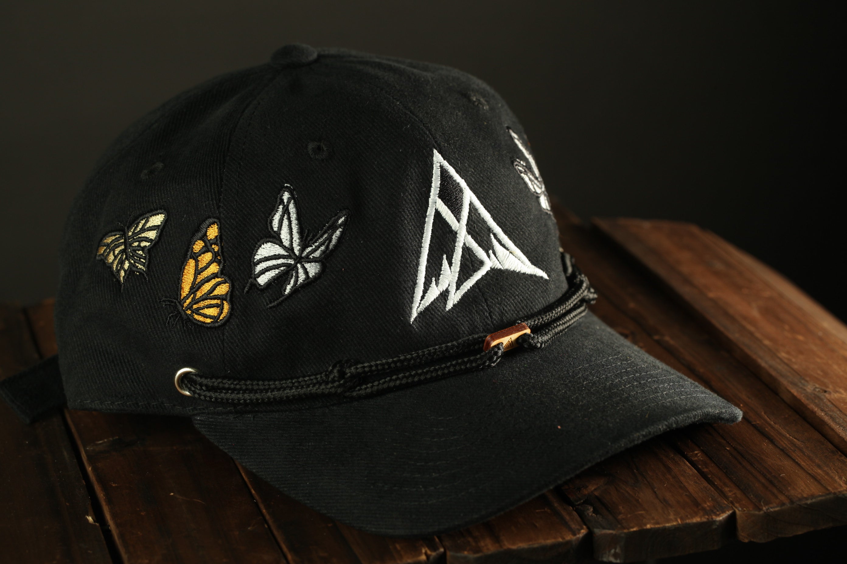 A black baseball cap with a white triangle design and three butterfly embroidered patches on the brim, resting on a wooden surface.