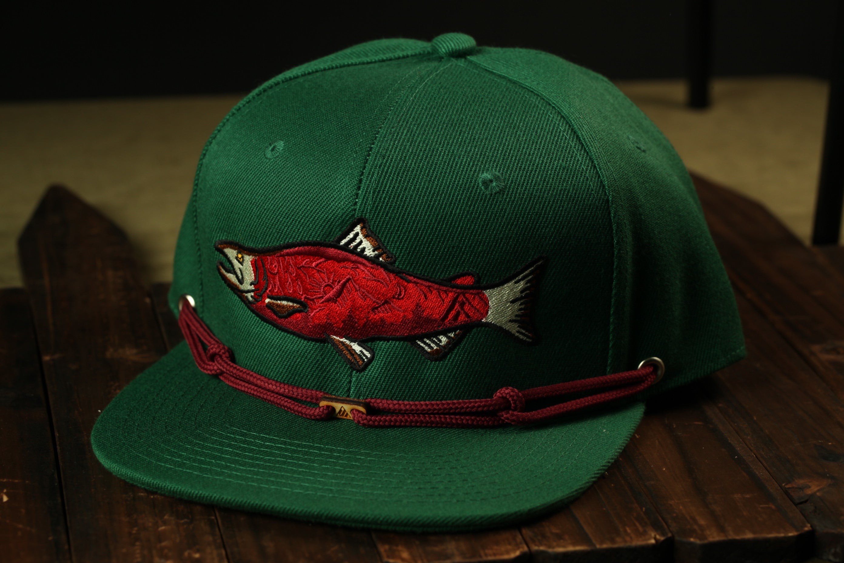 A green baseball cap with a red fish embroidered on it, resting on a wooden surface.
