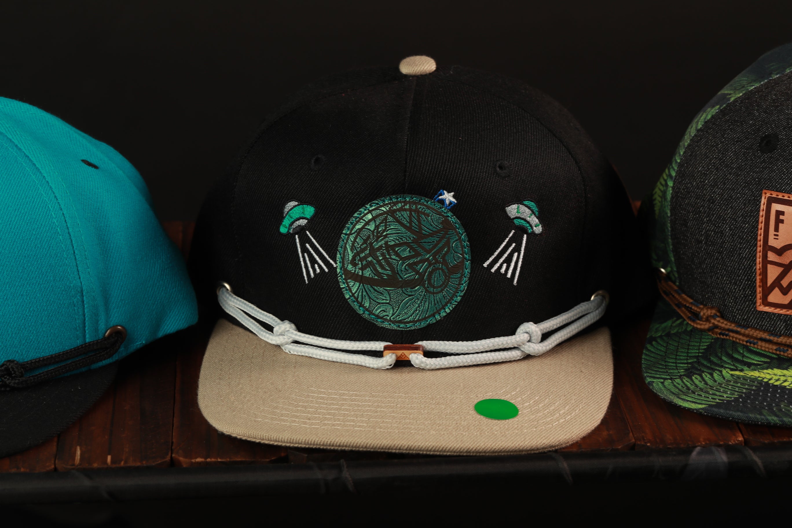 A black baseball cap with a green and white design, featuring a circular motif with a green alien figure and two white lines extending from the edges.