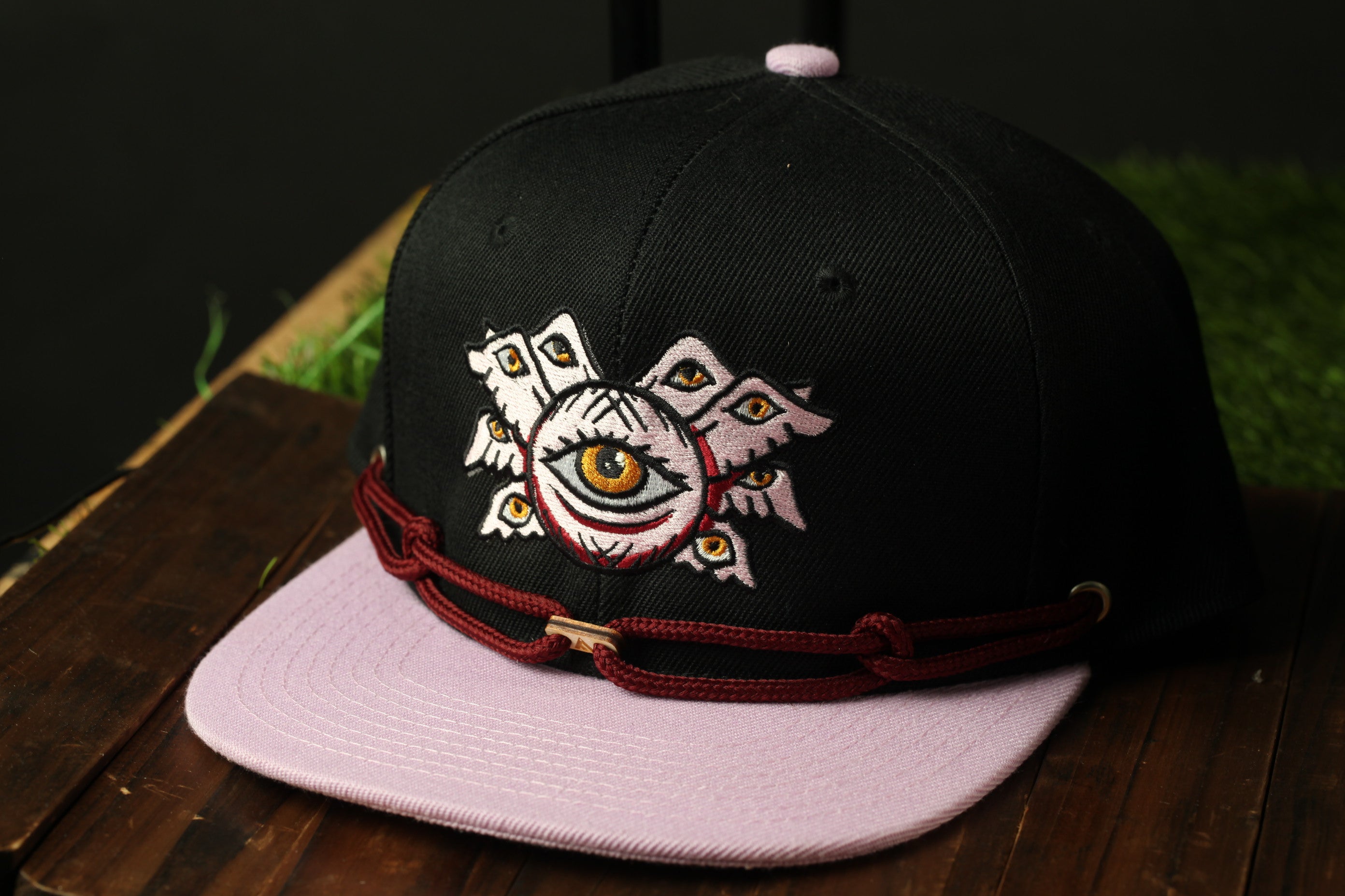 A black baseball cap with a pink brim and a white eye design on the front, sitting on a wooden surface.