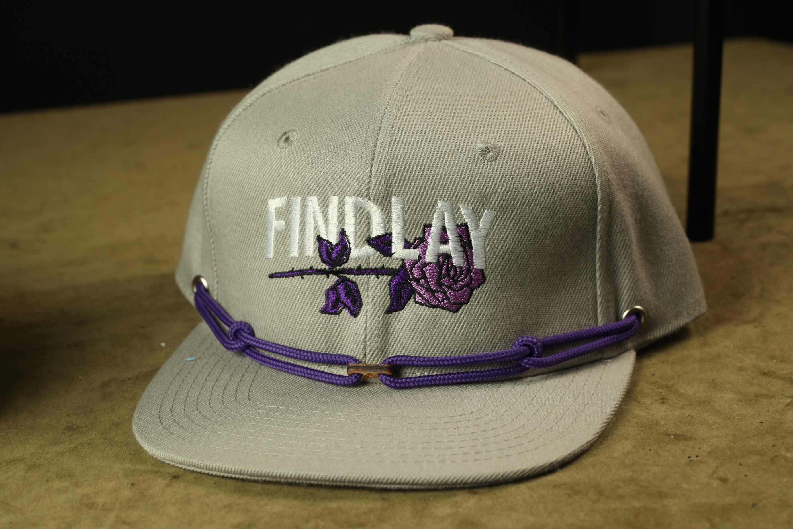 A gray baseball cap with a purple flower embroidered on it, and the word "FIND LAY" printed on the front.