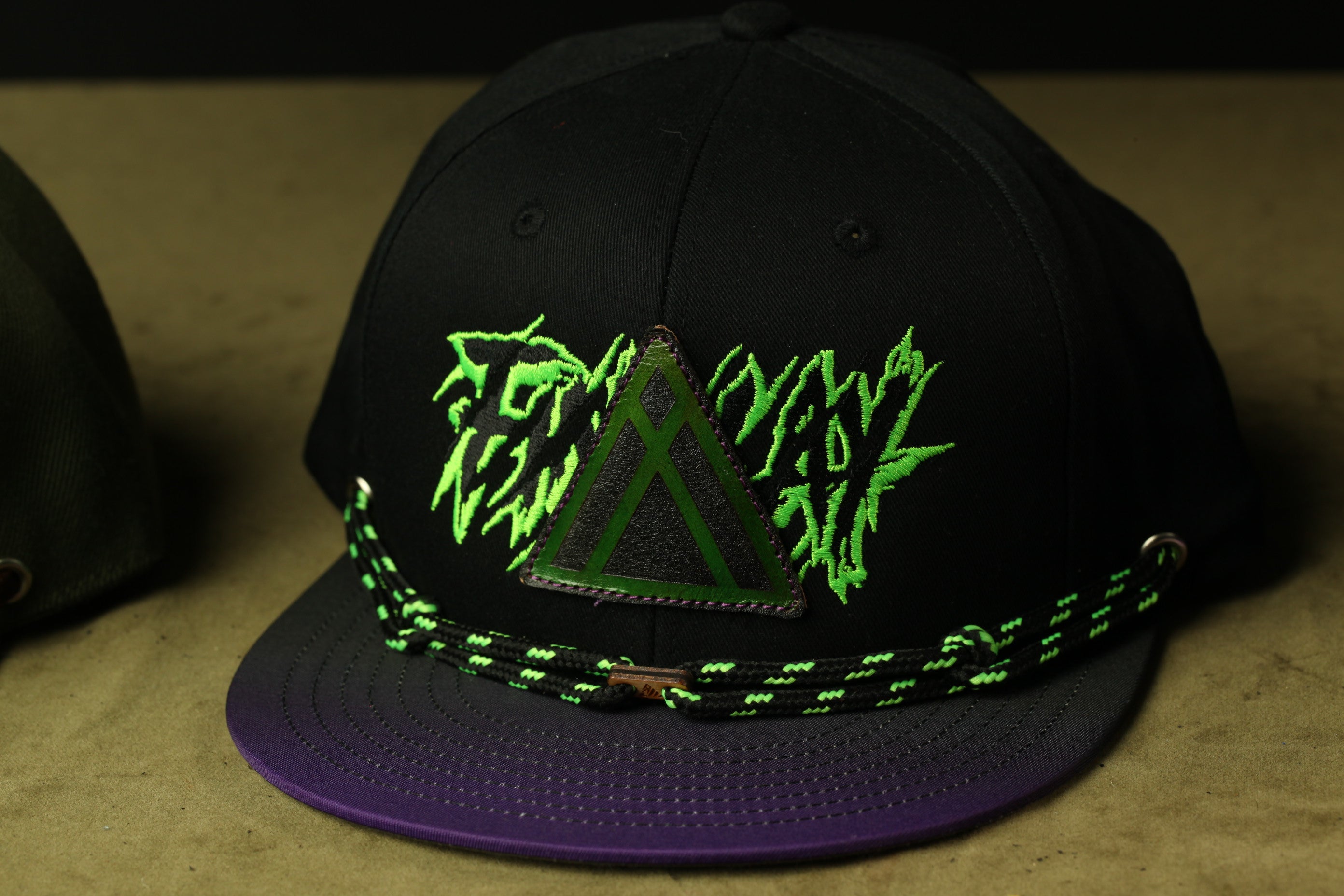 A black baseball cap with a purple brim and green and black embroidered designs.