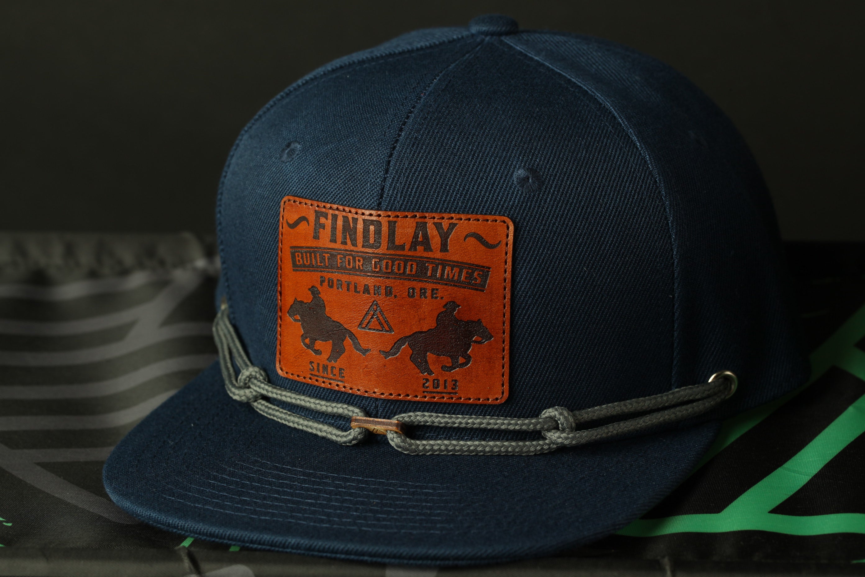 A navy blue baseball cap with a leather patch on the front, displaying the text "FINDLAY" and "BUILD FOR GOOD TIMES PORTLAND, OREGON SINCE 2013".